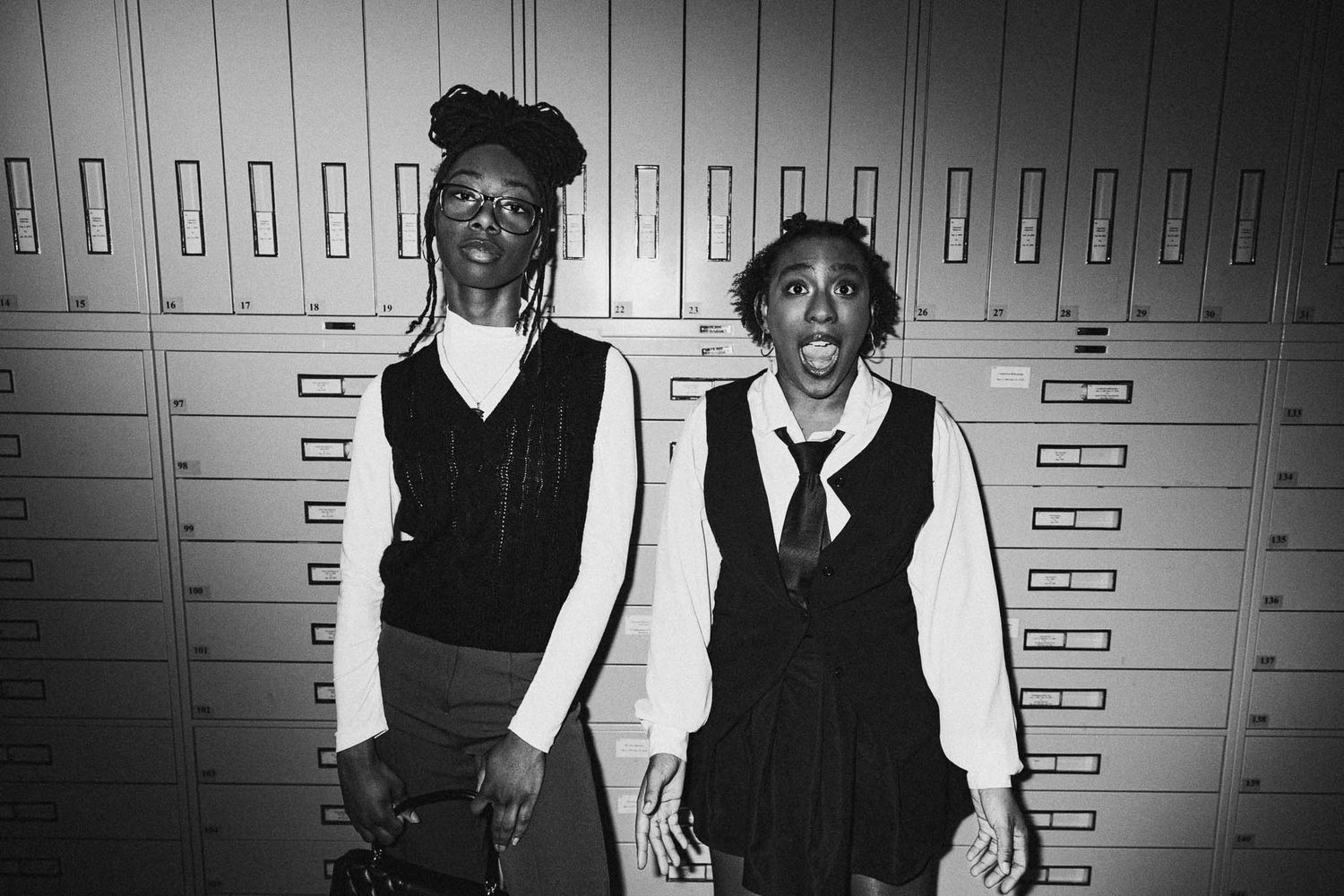 Two people pose expressively against a wall of lockers in black and white photography.