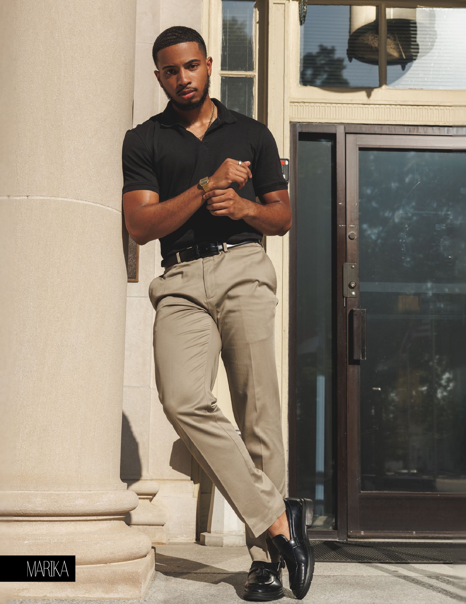 A stylish man in a black polo shirt and beige trousers leans against a white column in a sunlit outdoor setting.