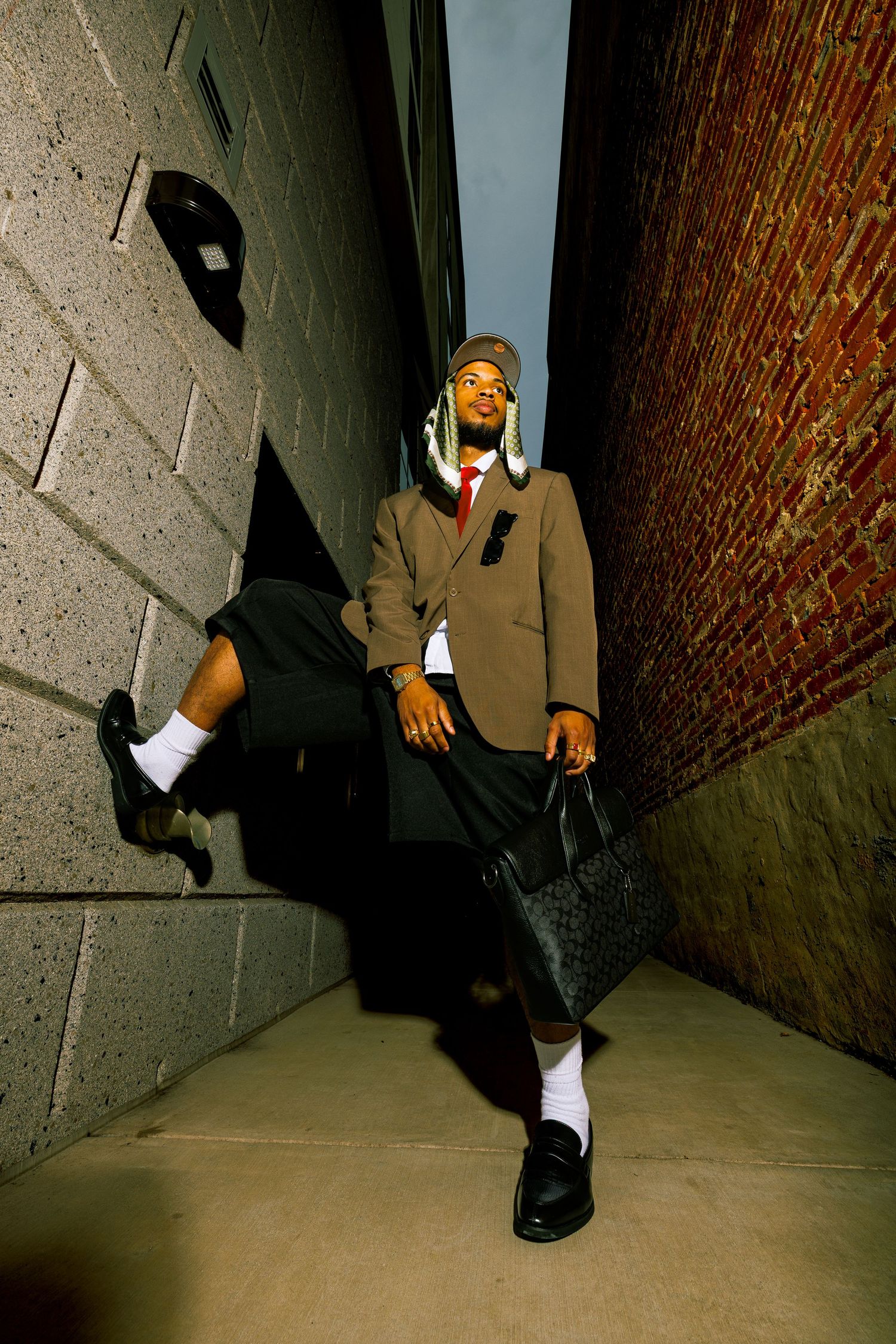 A stylish young Black man poses in an alley wearing a brown blazer, skirt, hat, white socks, and loafers while holding a bag.