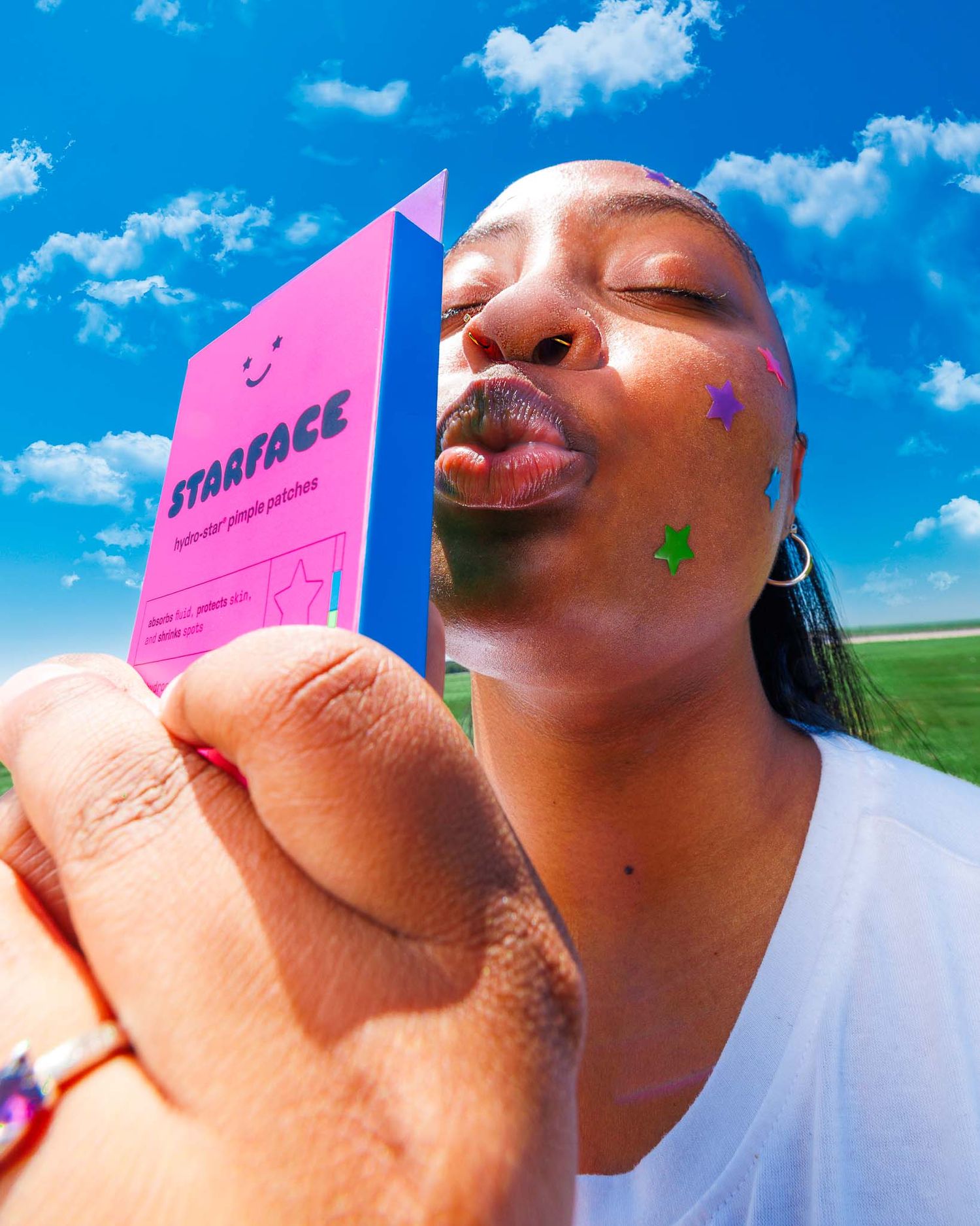 Woman holding Starface hydro-star pimple patches box outdoors with colorful star stickers on her face.