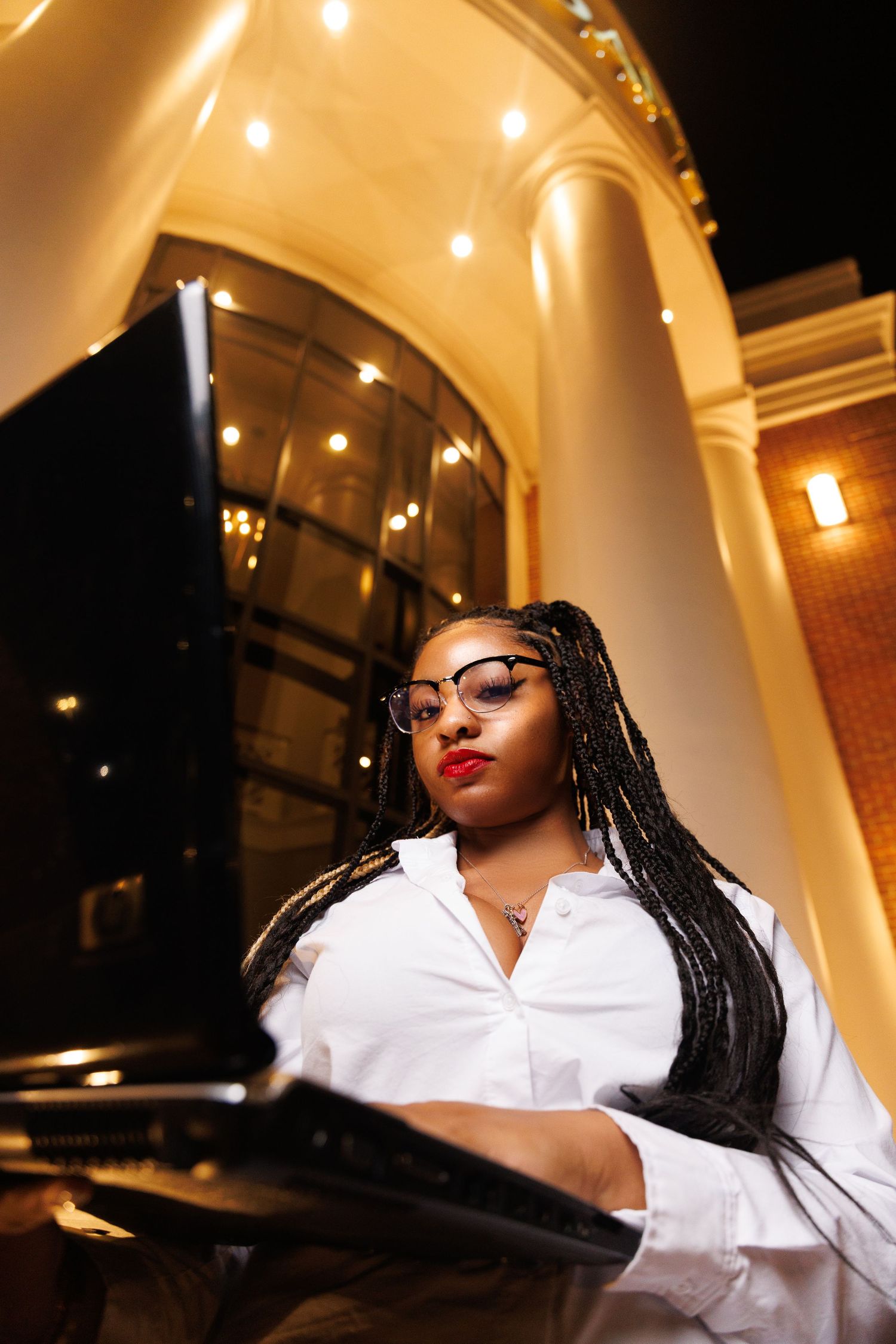 A young Black woman with braids and glasses works on a laptop outside an elegant building at night.