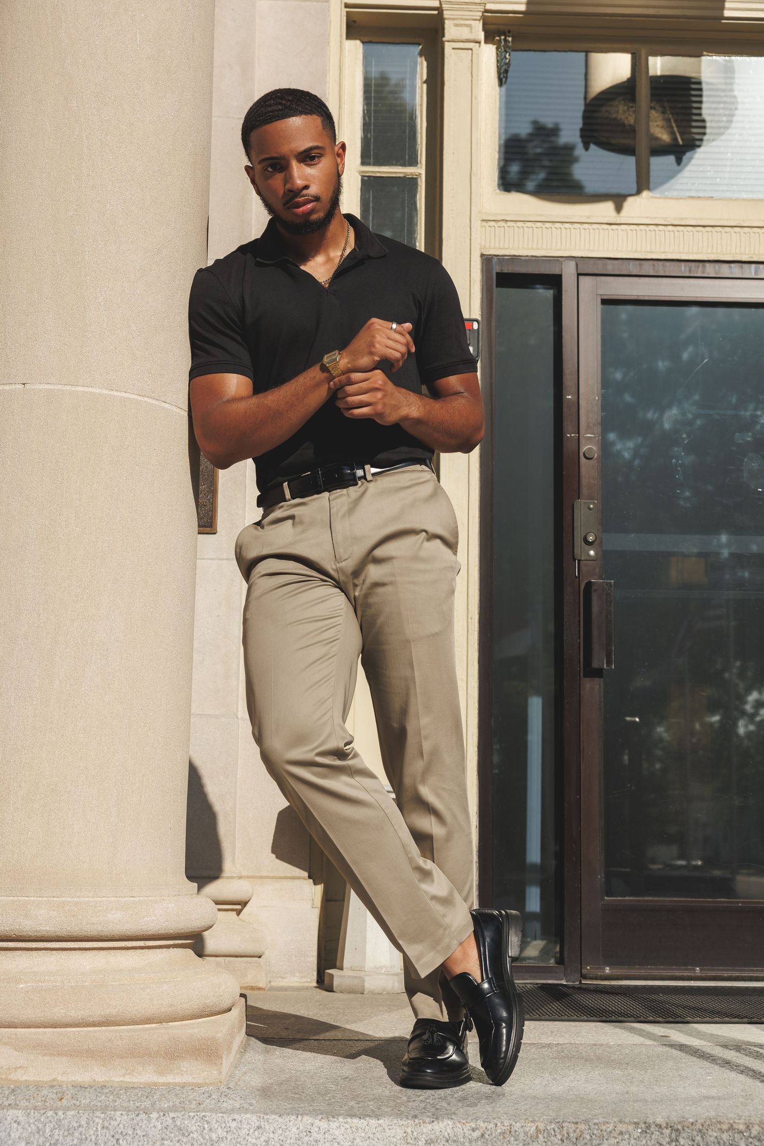 A stylish young Black man in a black polo shirt and beige trousers leans against a column outdoors.