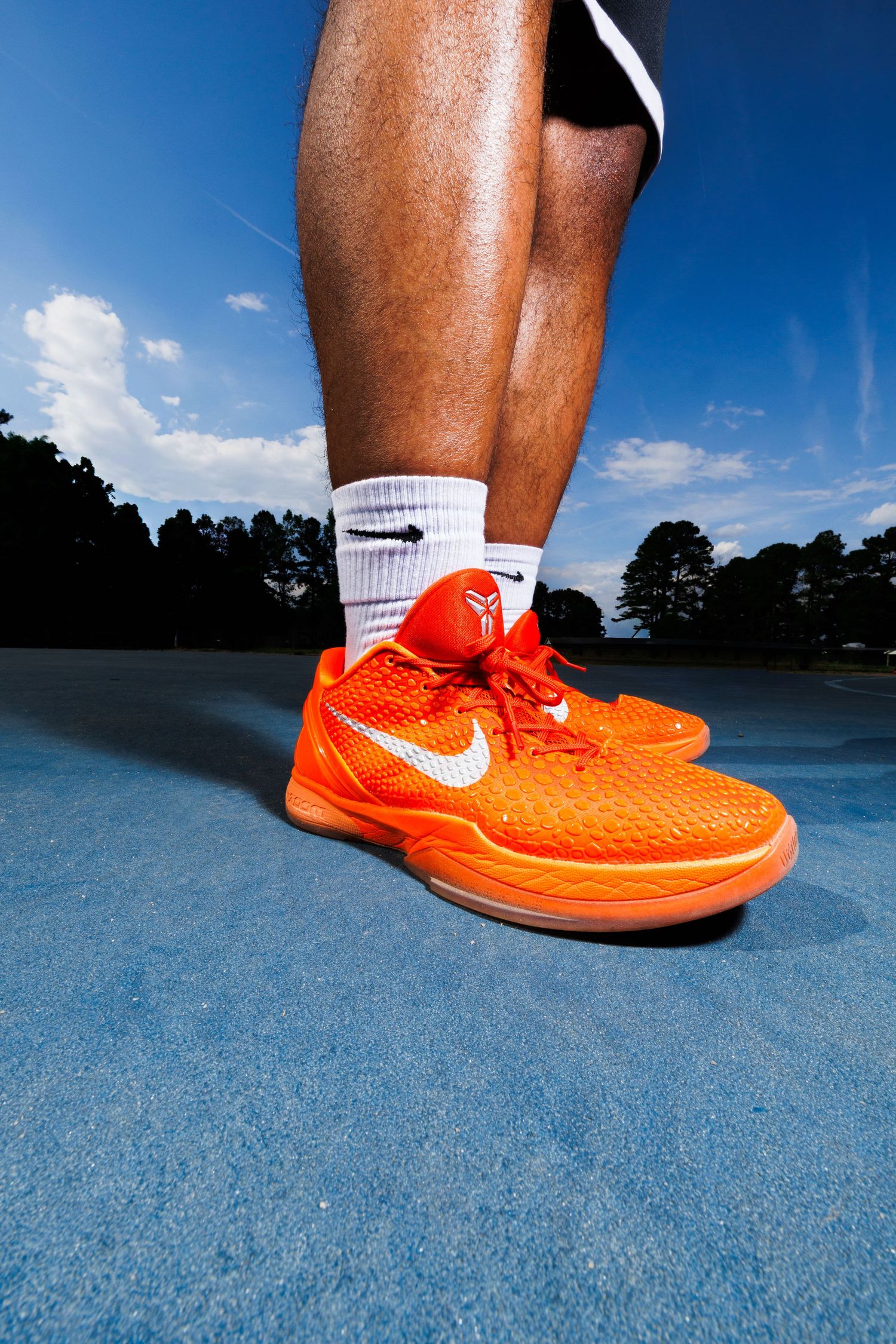 Person wearing orange Nike Kobe basketball shoes with white socks on a blue court outdoors.
