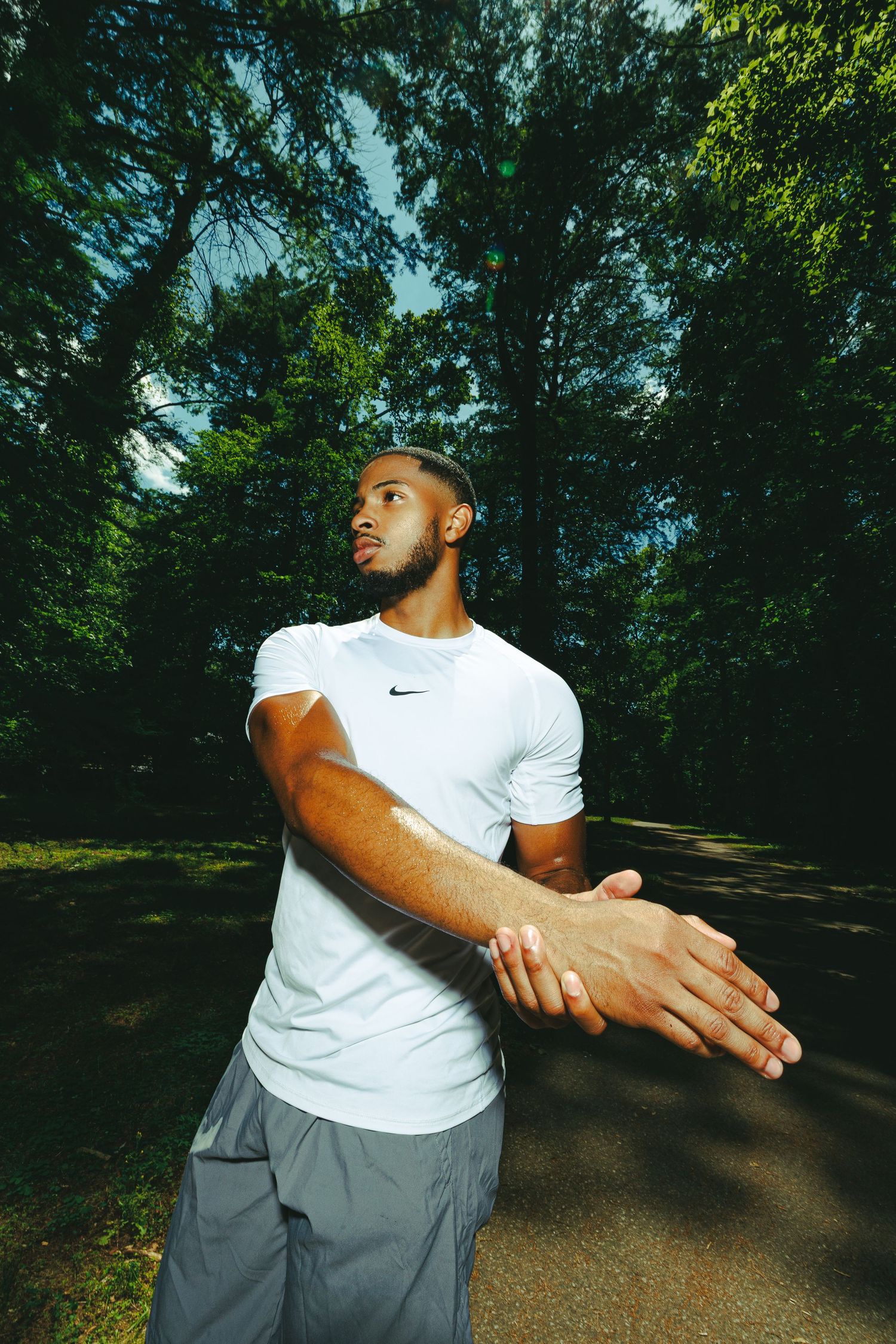 Athletic man in white Nike shirt stretching arms outdoors on a tree-lined path before a run.