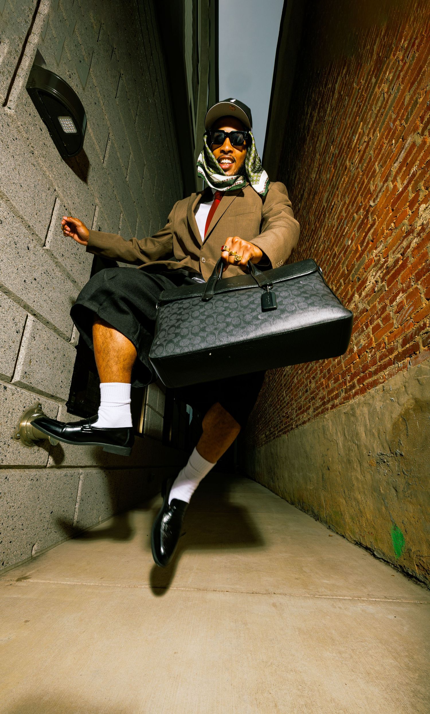 Man in eclectic outfit with sunglasses, cap, blazer, shorts, and loafers holds large designer bag in urban alley.