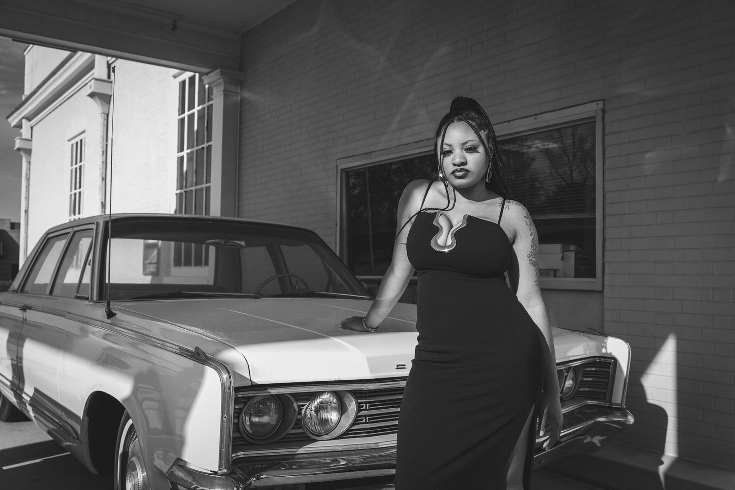 Black and white photo of a woman in a sleek dress posing beside a vintage classic car in a rustic garage setting.
