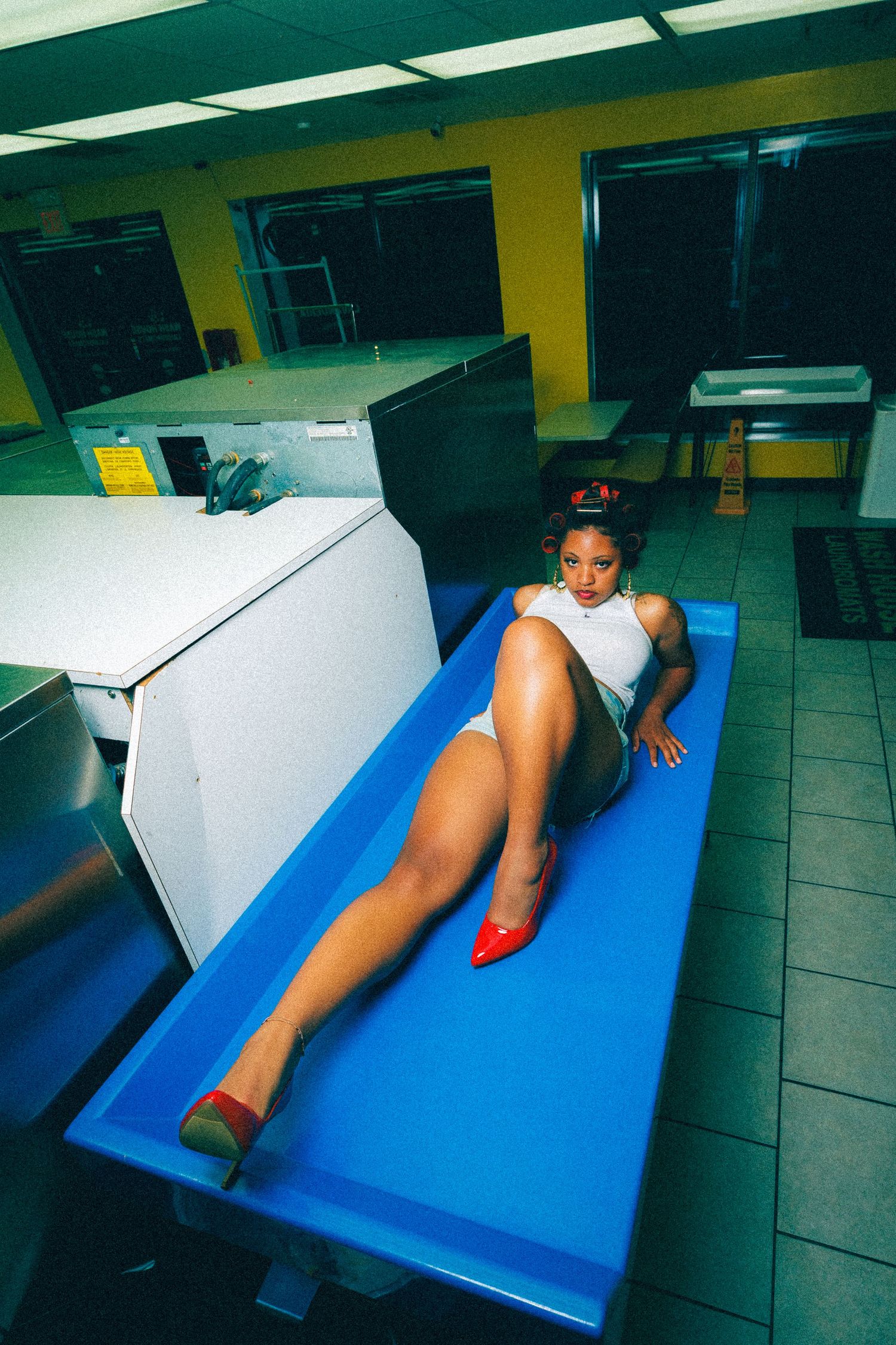 Woman in white outfit and red heels posing on a blue table in a laundromat setting.