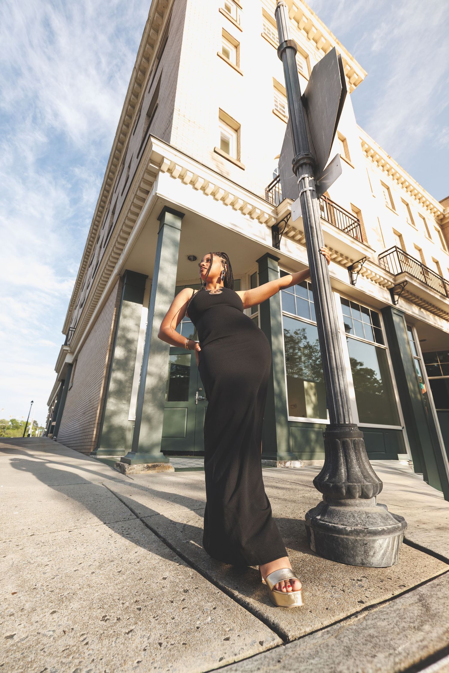 Pregnant woman in black maxi dress poses by street lamp outside historic building on sunny day.