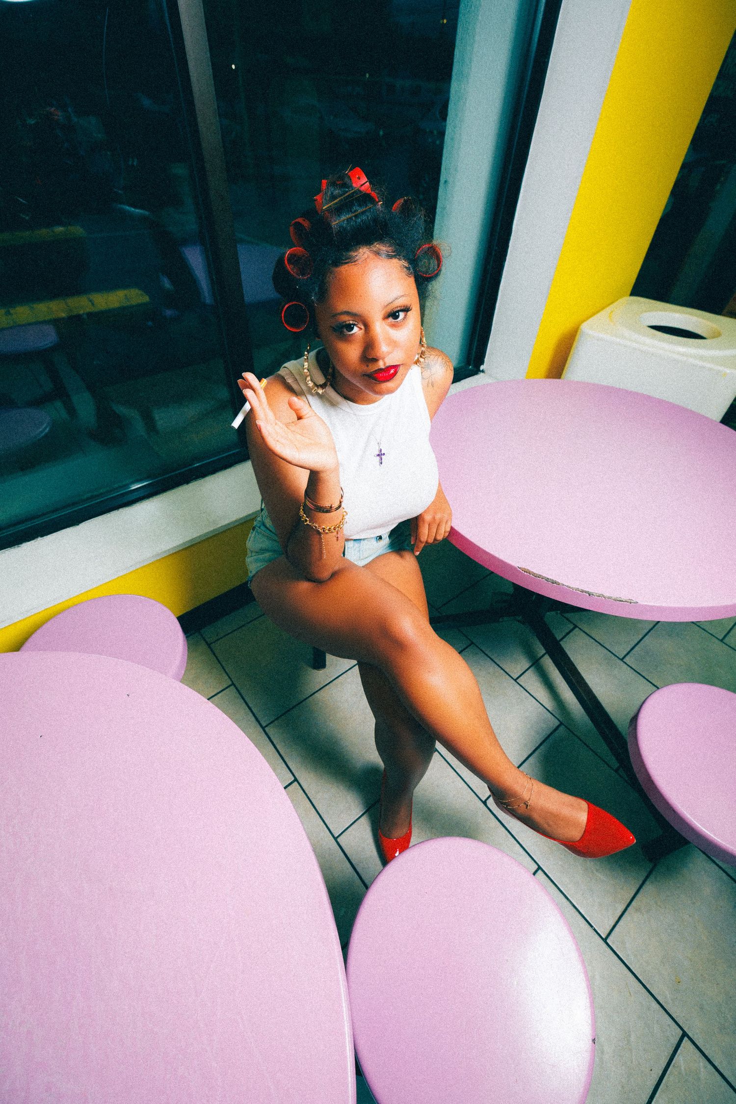 Young woman with red hair rollers sits confidently at pink tables in a colorful diner, wearing white top and red heels.