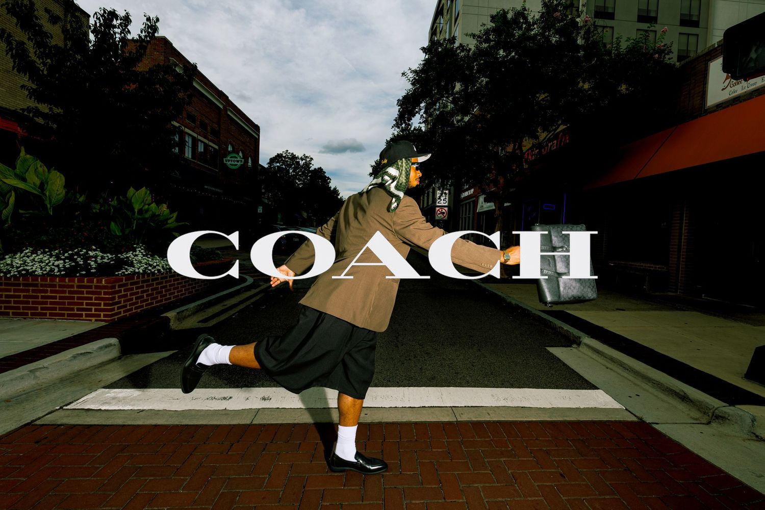 Person crossing a street in a Coach fashion campaign, wearing a brown oversized coat and white socks.