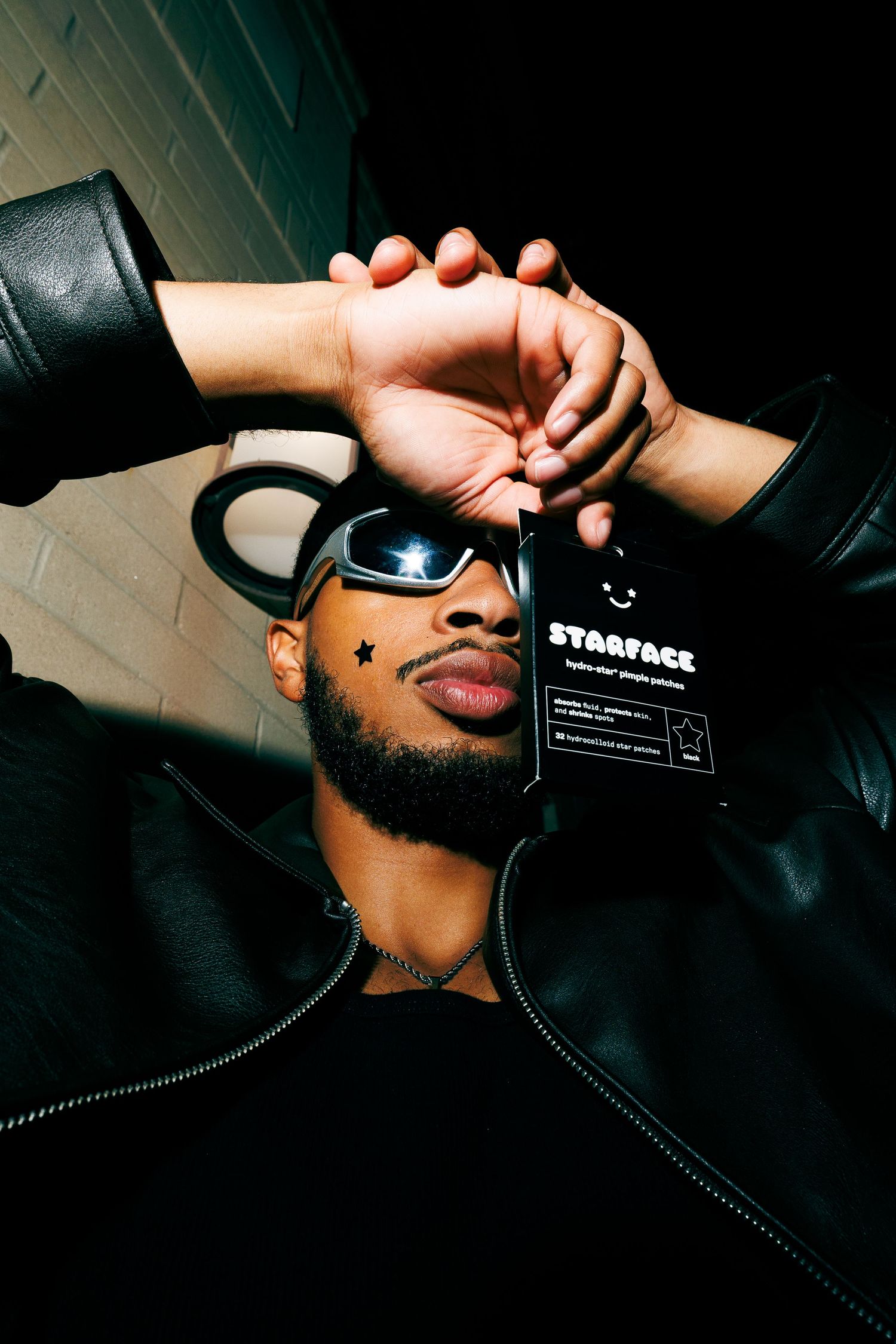 Man in black leather jacket and reflective goggles holds Starface skincare product in dramatic low-light photo.