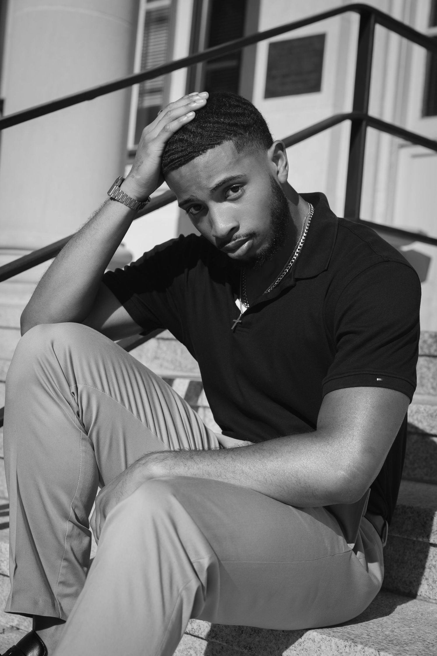 Black and white portrait of a young man sitting on steps, hand on head, wearing a polo shirt and chain necklace.