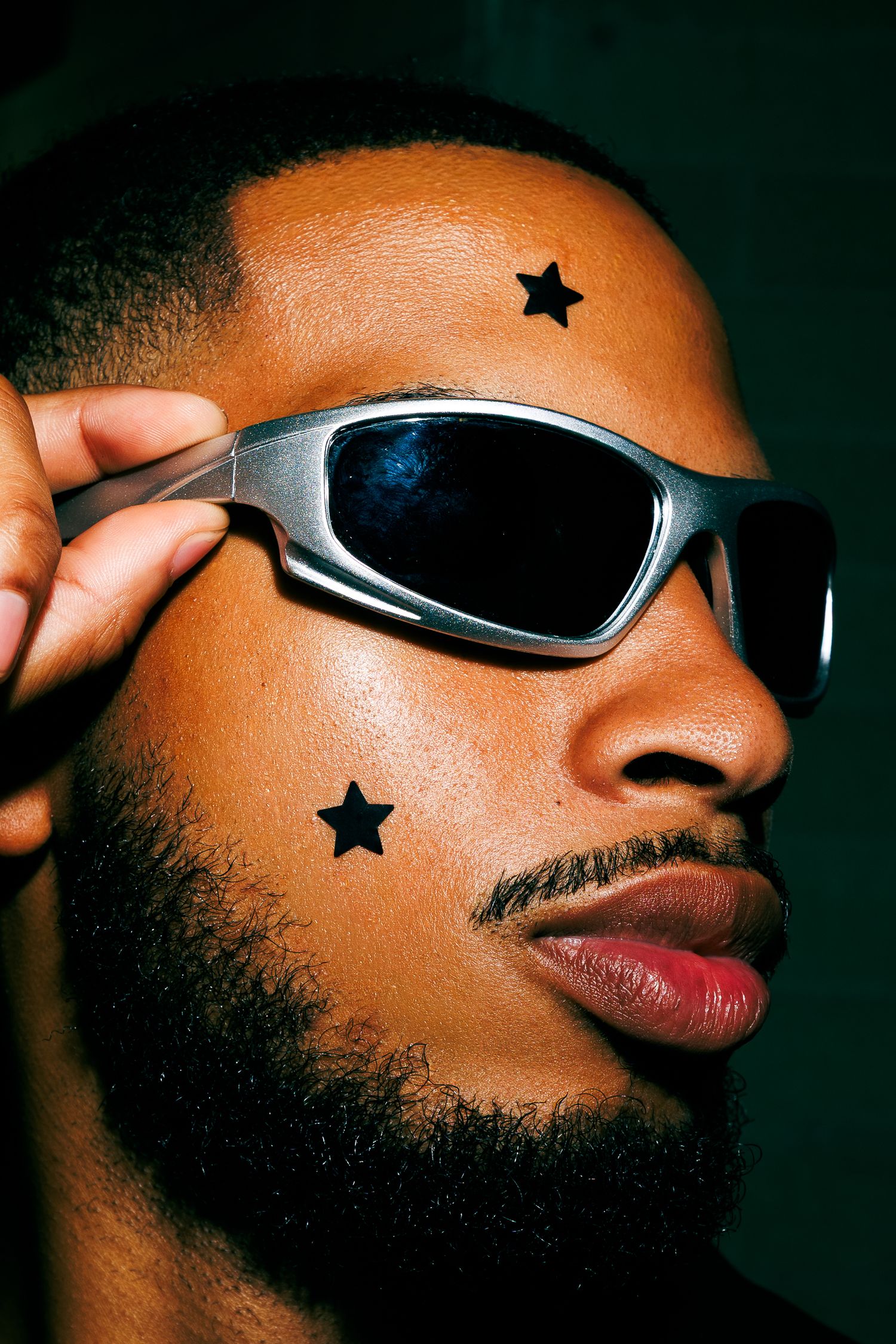 Close-up of a man with star face tattoos adjusting silver wraparound sunglasses against dark background.
