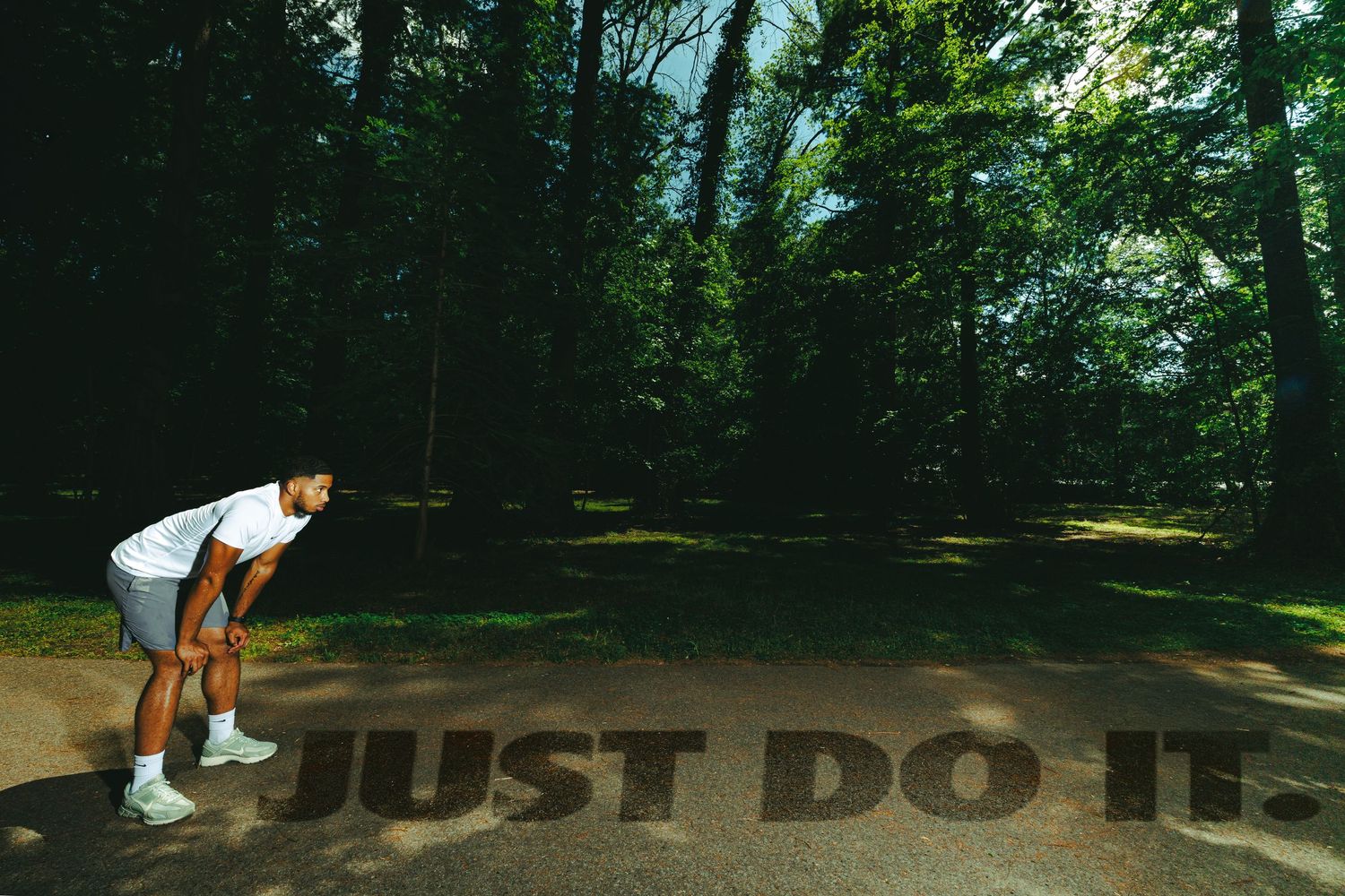 Athlete in white sportswear bends over Nike's 'Just Do It' slogan on a path surrounded by lush green trees.