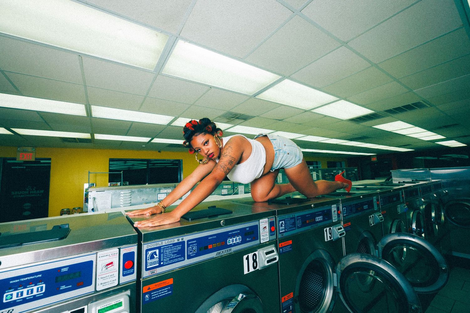 Woman posing on top of washing machines in a colorful laundromat setting.