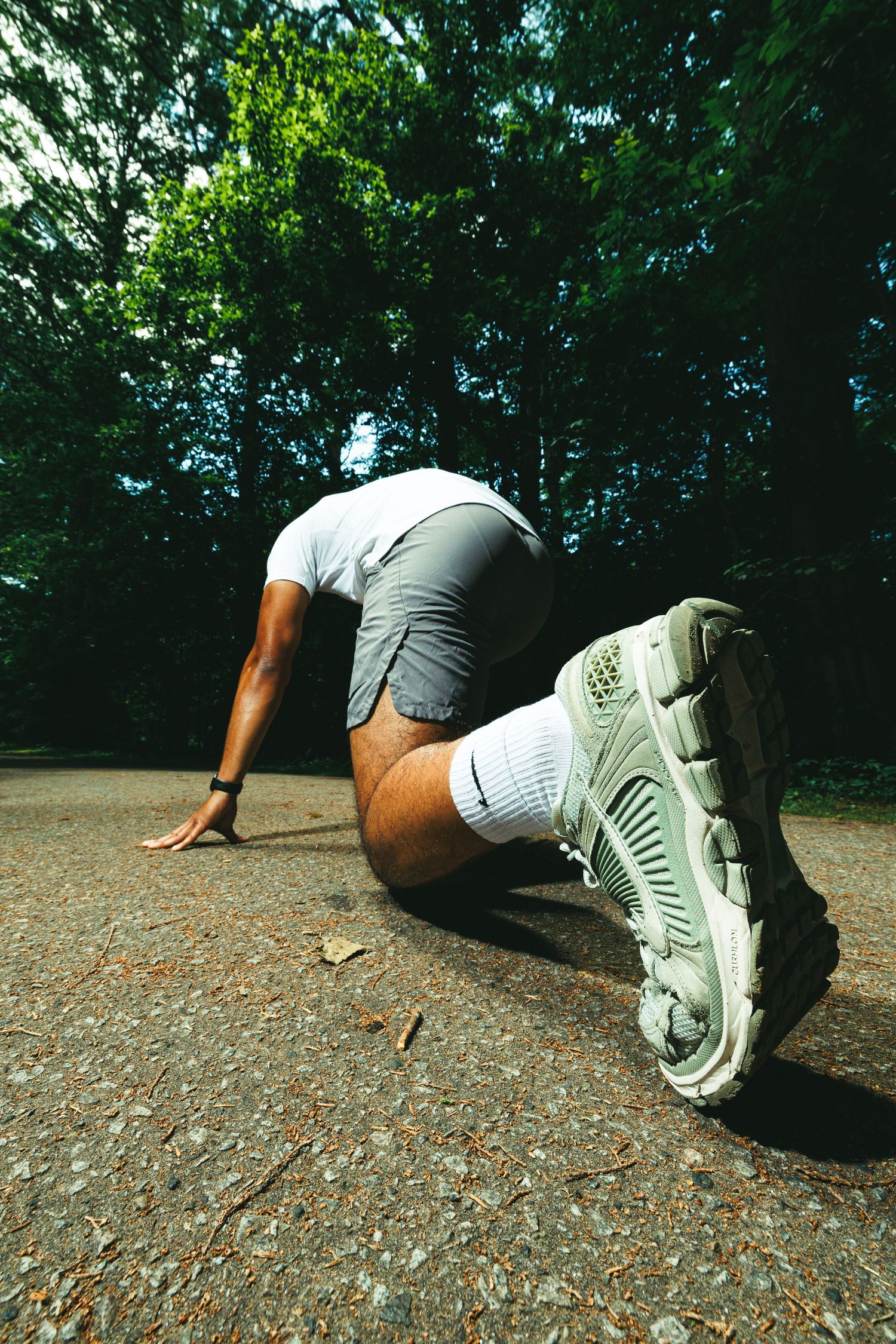 A runner in gray shorts and white sneakers crouches in a starting position on a path surrounded by green trees.