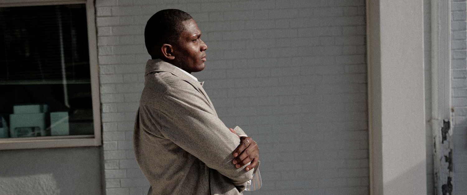 Person in beige coat standing against gray wall looking contemplative.