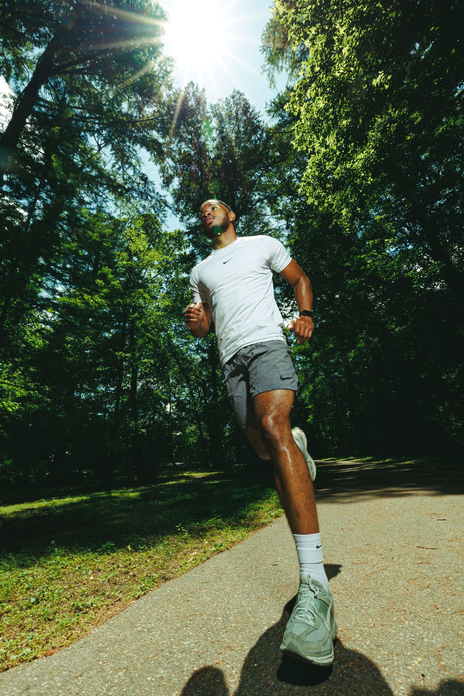 A man in Nike athletic wear jogs on a sunlit park path surrounded by lush green trees on a bright sunny day.