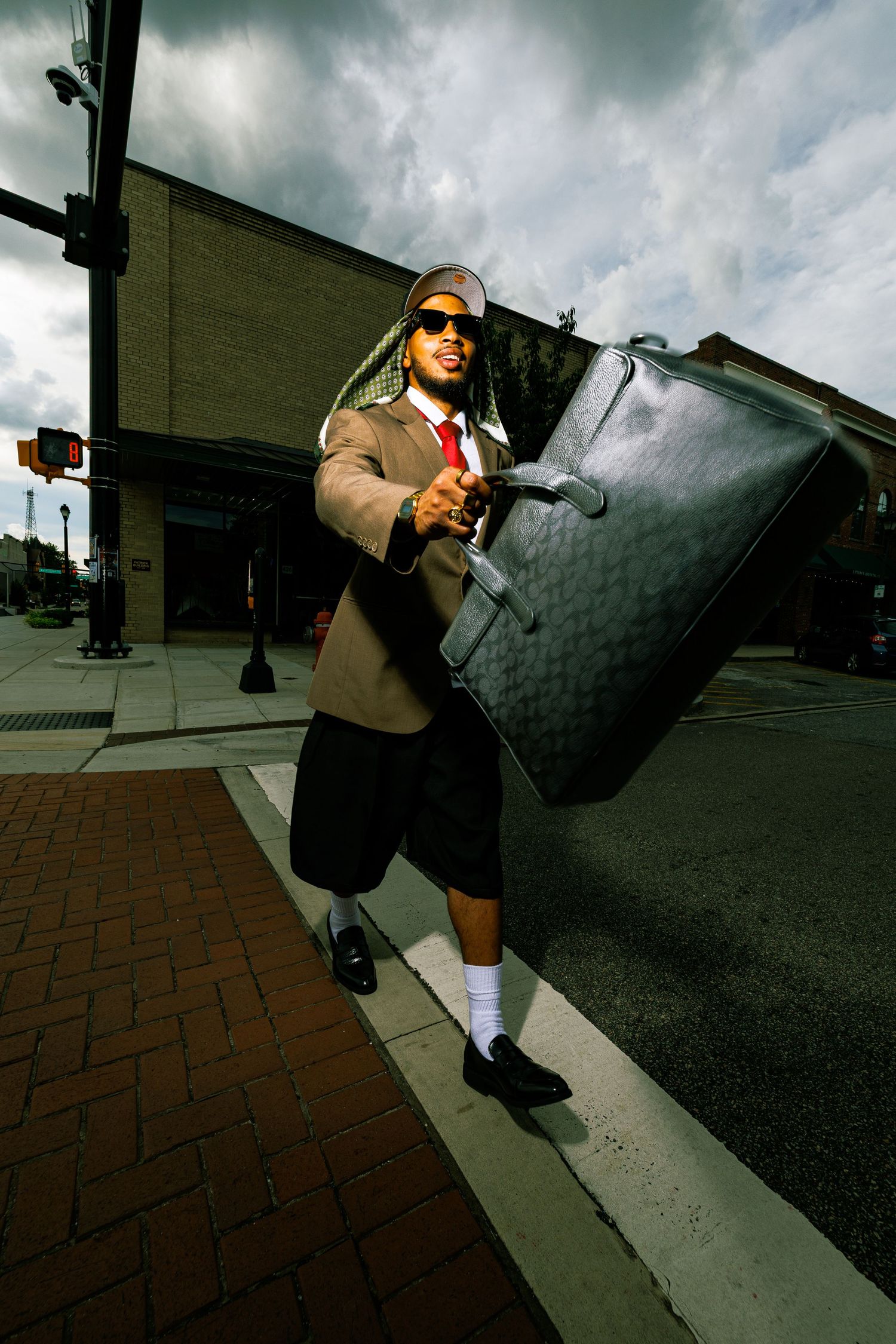 Stylish man with braids, sunglasses, blazer, shorts, holding large designer bag on urban street corner.