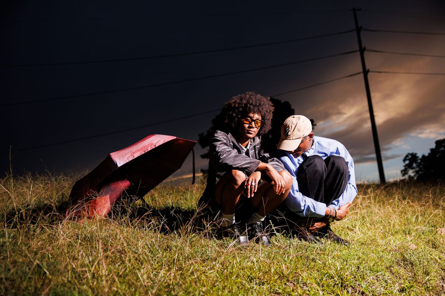 Two people sit together in a grassy field at dusk under a dramatic stormy sky near power lines.