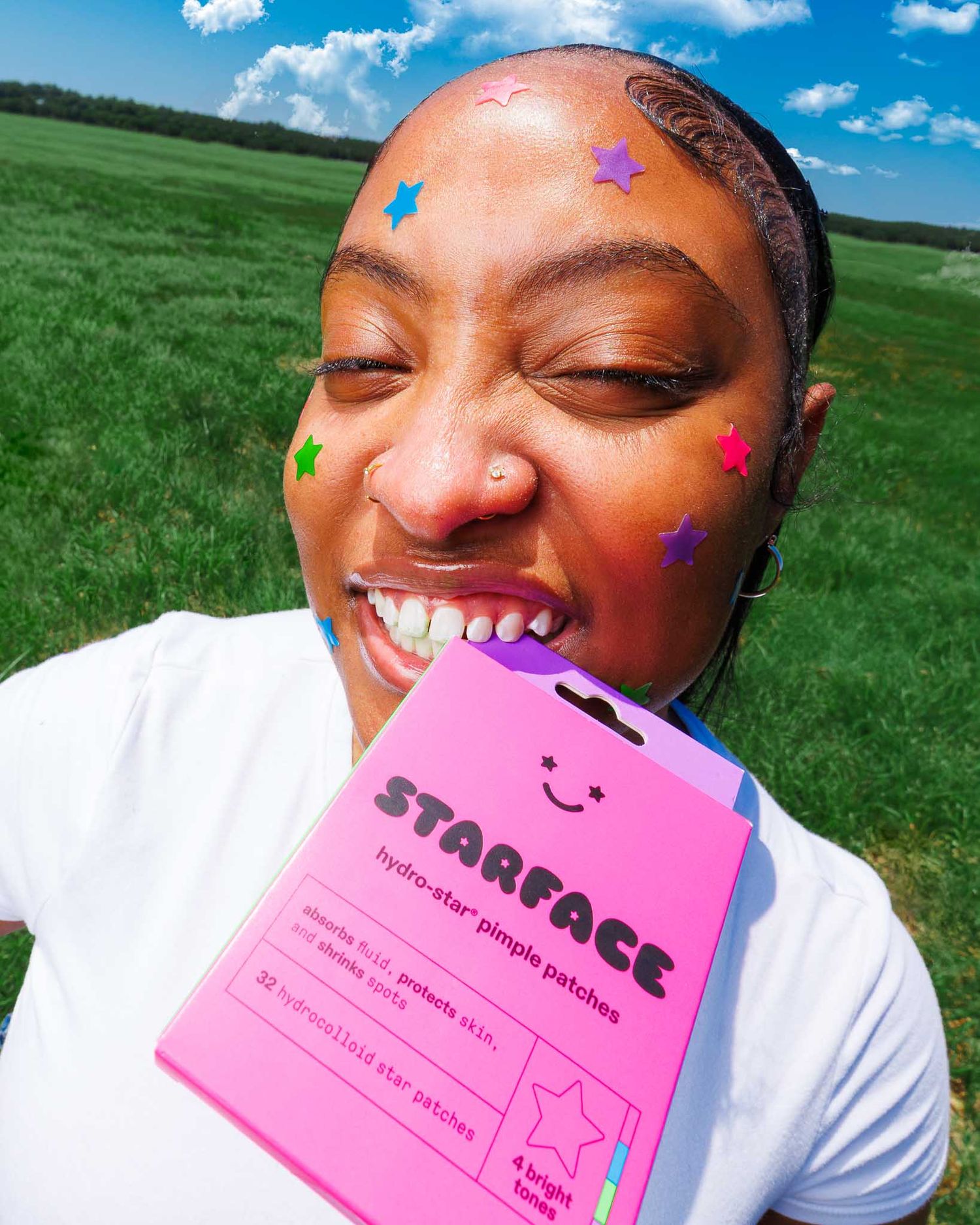 Young woman smiling outdoors holding Starface pimple patches box with colorful star stickers on her face.