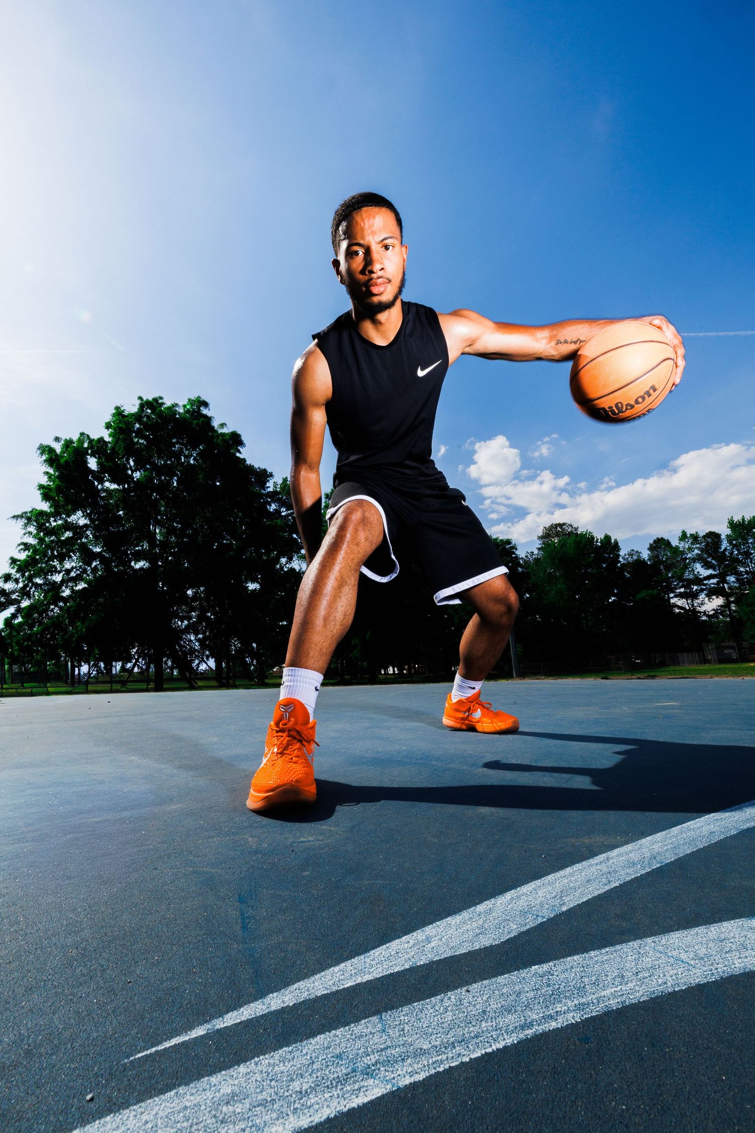 Athletic man in black Nike outfit dribbling basketball on outdoor court with blue sky background.