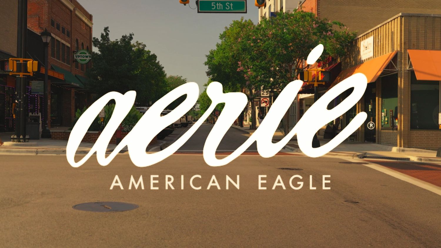 Aerie by American Eagle logo overlaid on a sunny small-town street scene with shops and trees.
