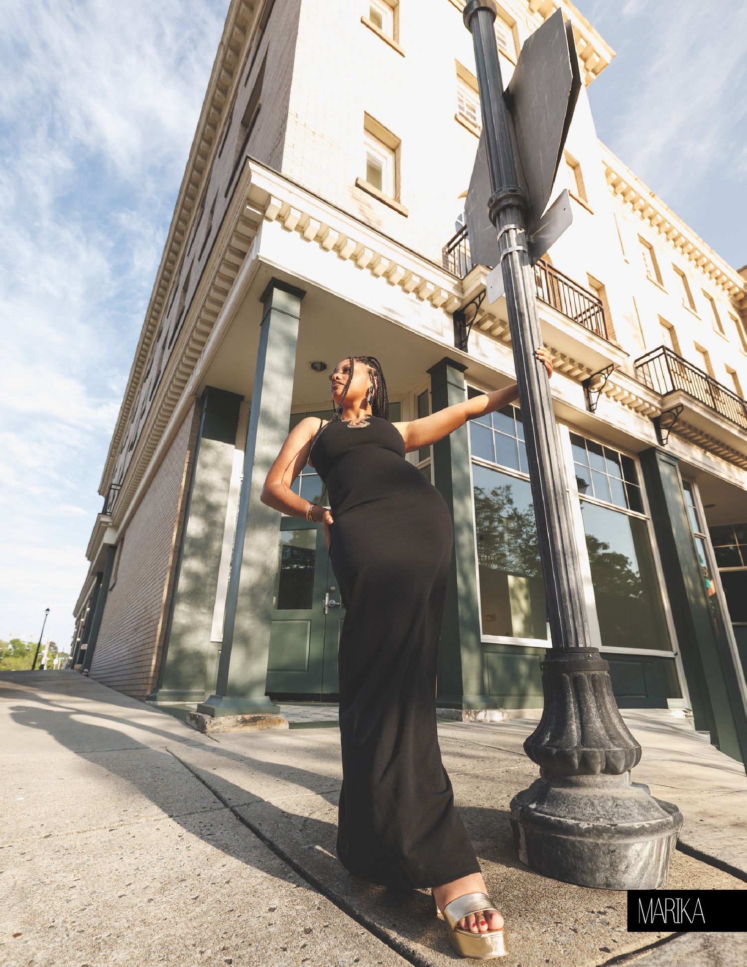 Woman in black maxi dress poses by street lamp outside historic building on sunny day.
