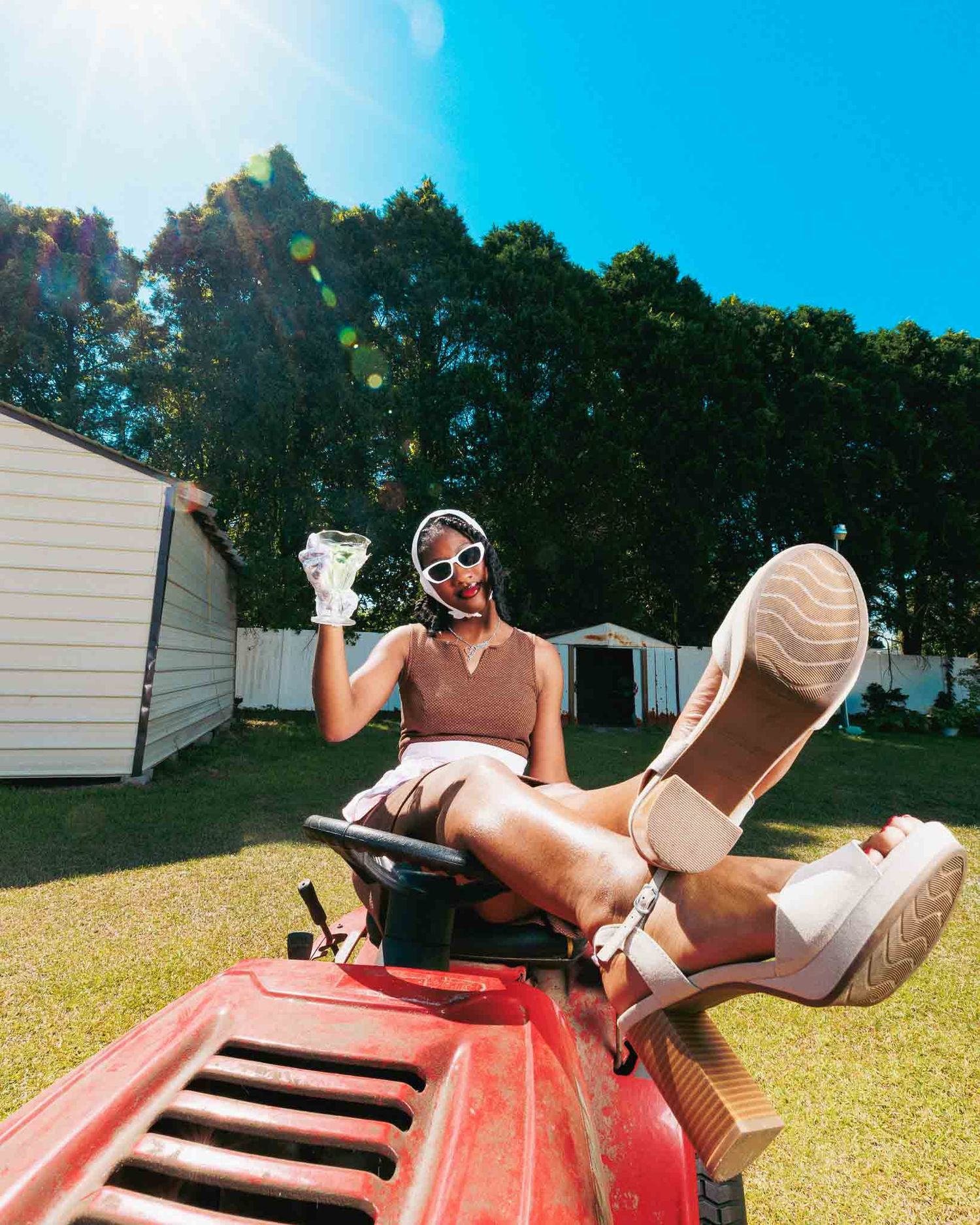 Woman relaxing on red ride-on mower, holding drink, wearing sunglasses, white shorts, on sunny green lawn.