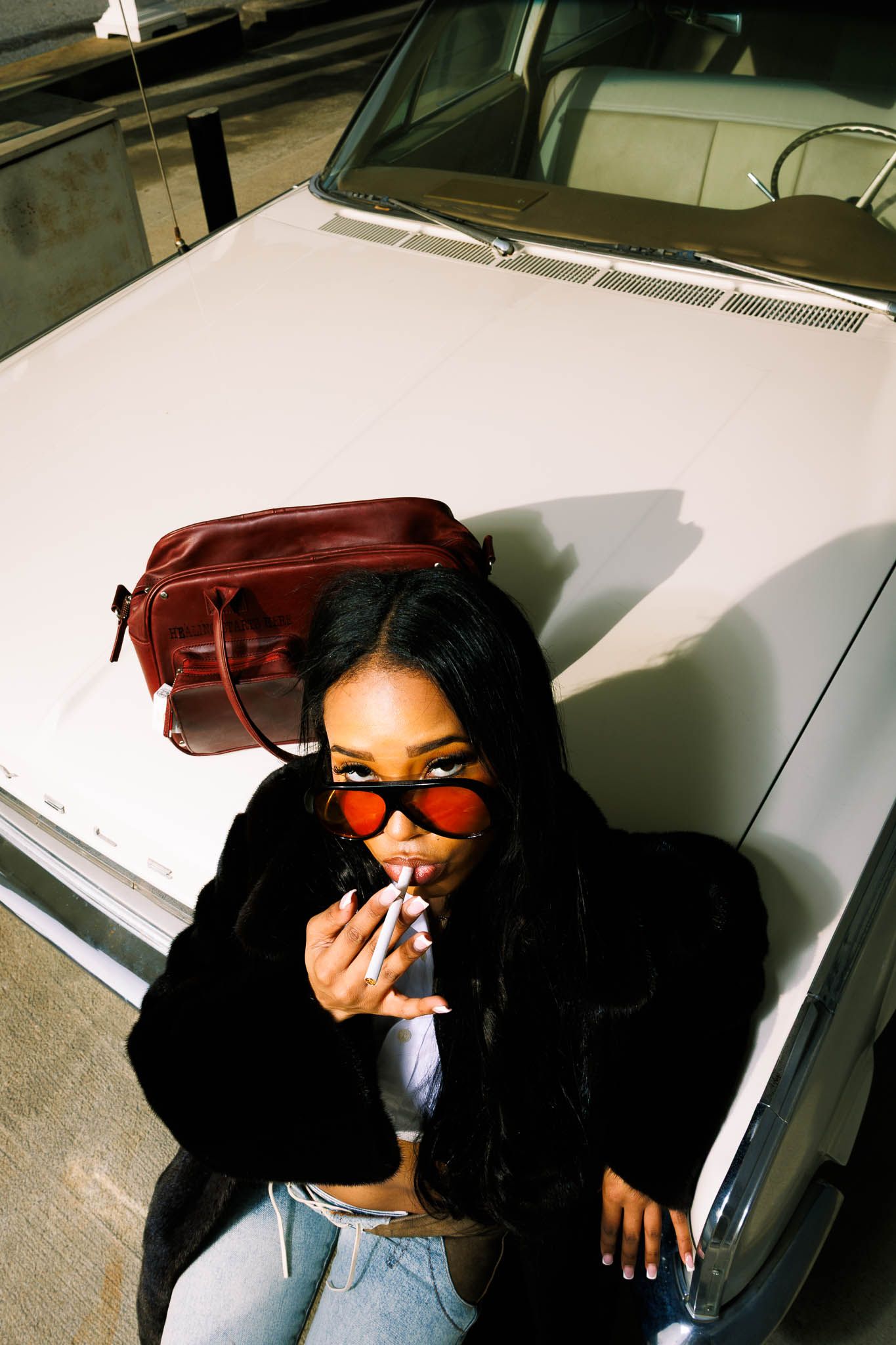 Figure in black clothing and red sunglasses posing against white car.