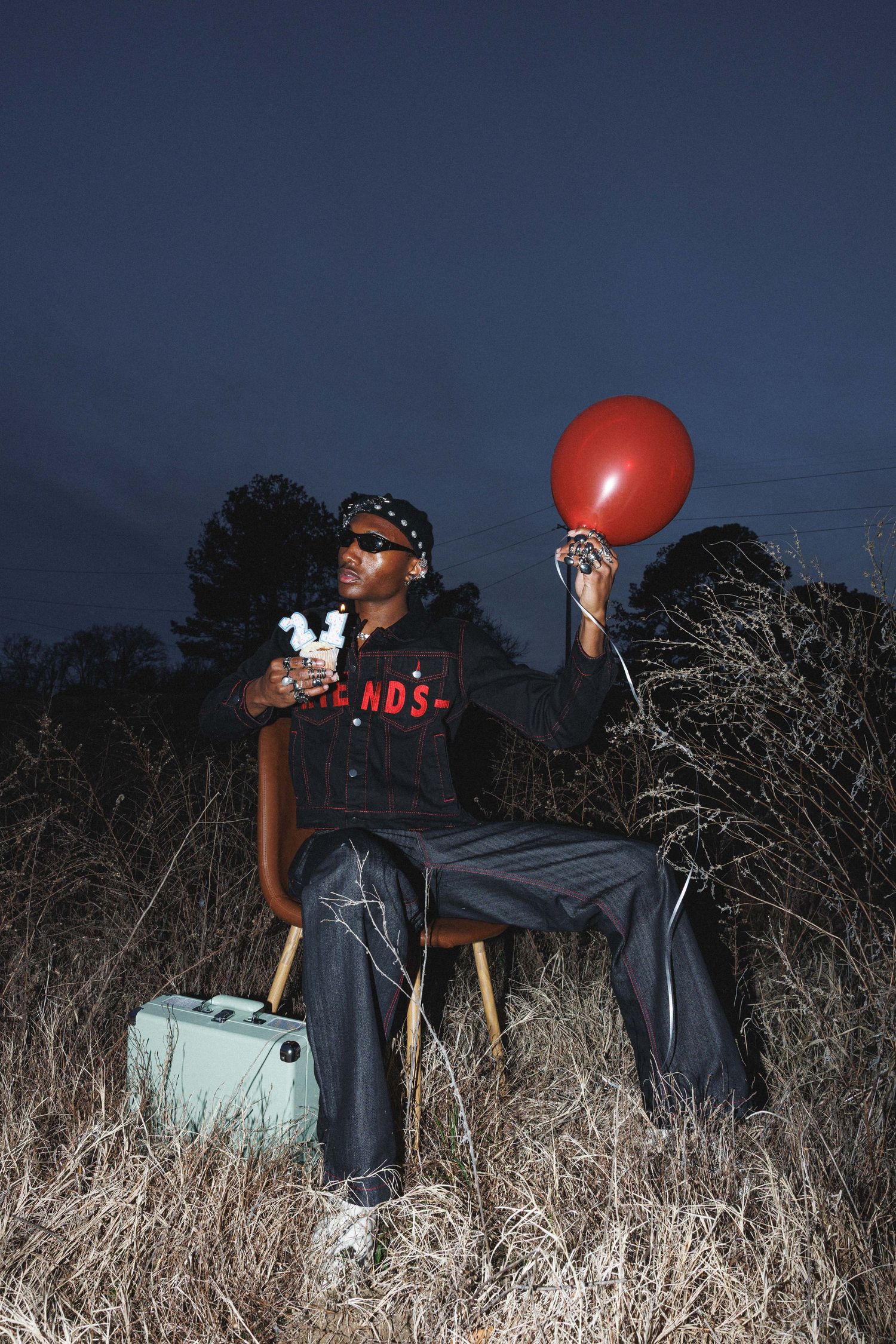 Conceptual editorial portrait featuring a model holding a cupcake and a red balloon in an open field during blue hour.