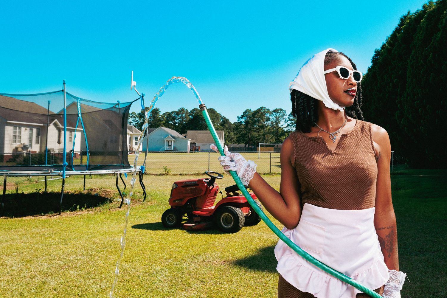 Woman in sunglasses and white hat holds garden hose in sunny backyard with trampoline and red lawn mower.