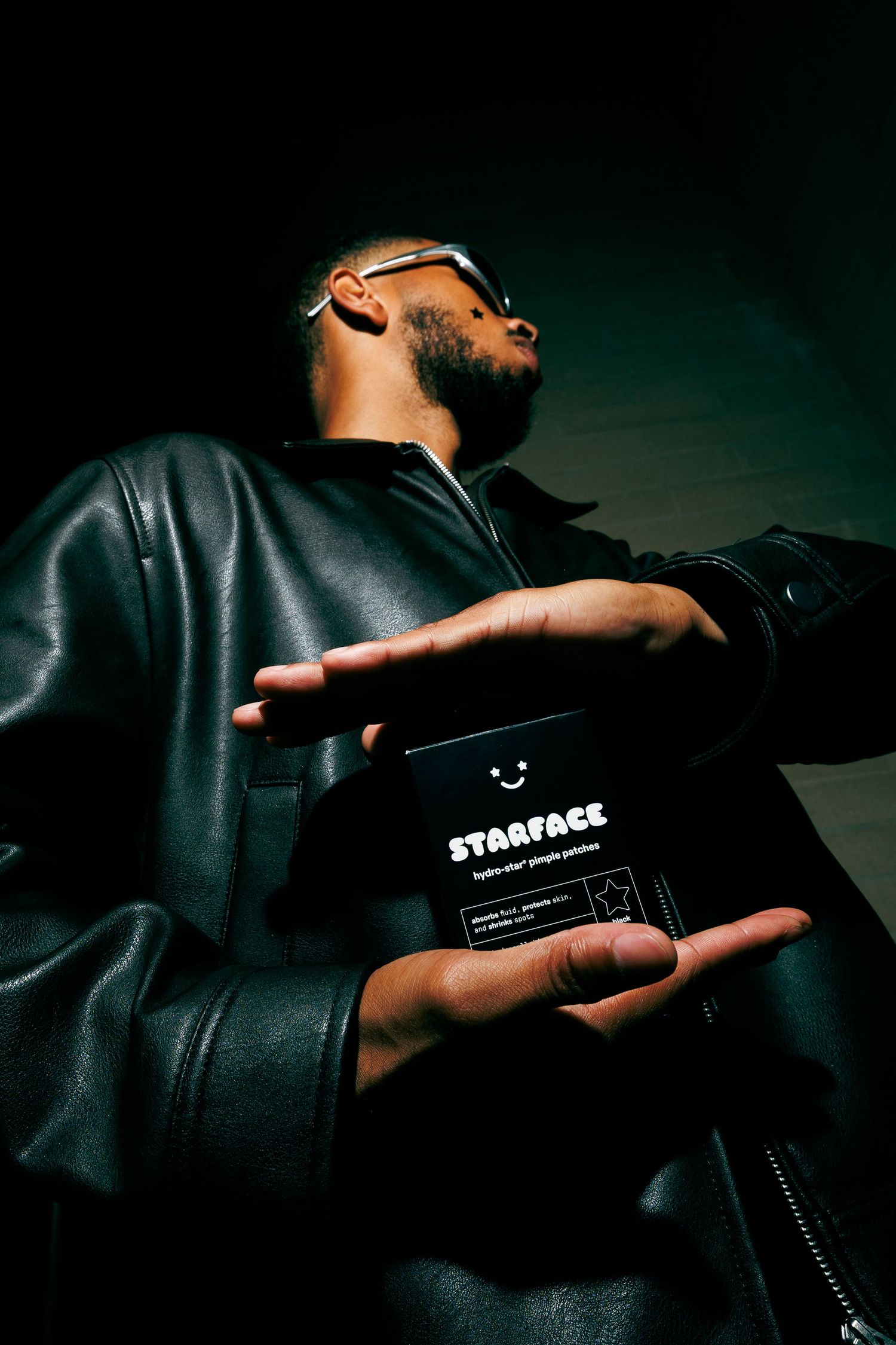 Man in black leather jacket holding Starface hydrocolloid pimple patches box while wearing silver sunglasses.