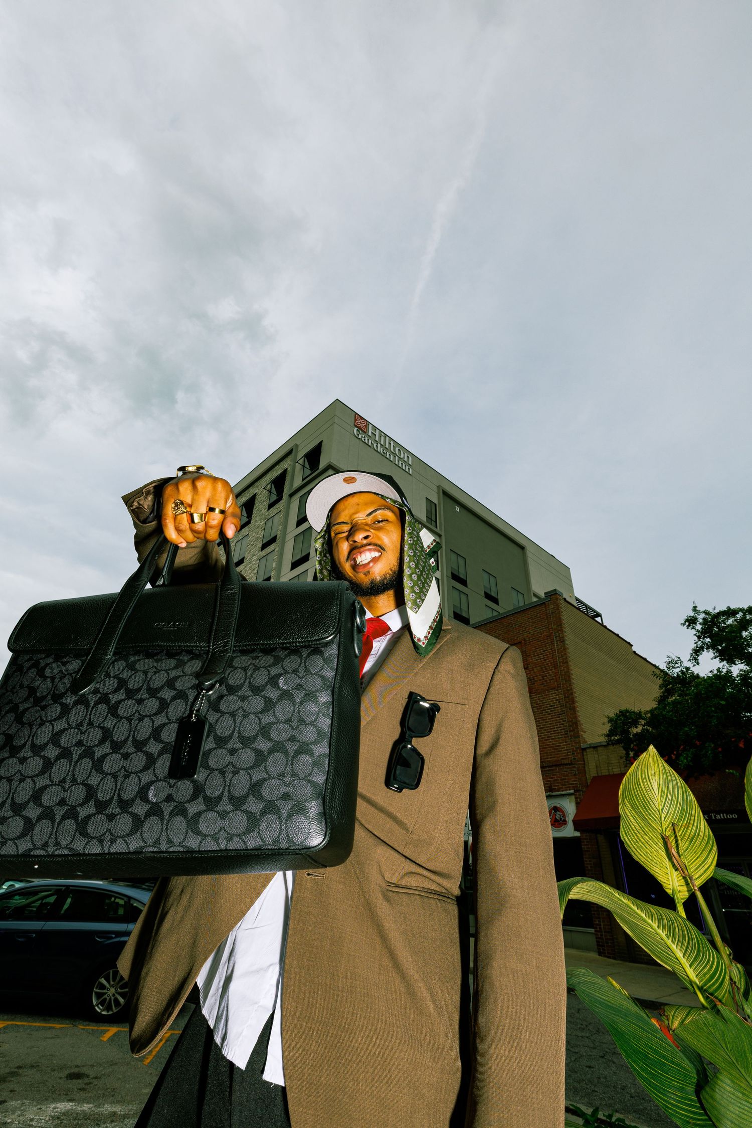 Stylish man in brown suit holding a Coach briefcase outdoors near a hotel building on a cloudy day.