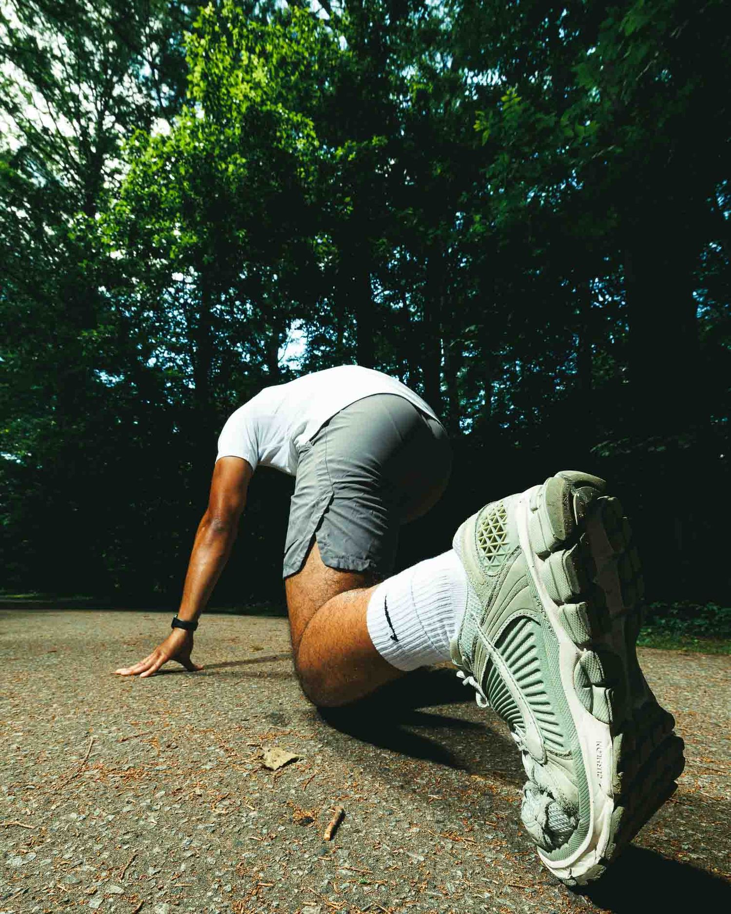 A runner crouches in a starting position on a park path, wearing gray shorts, white socks, and sneakers, ready to sprint.