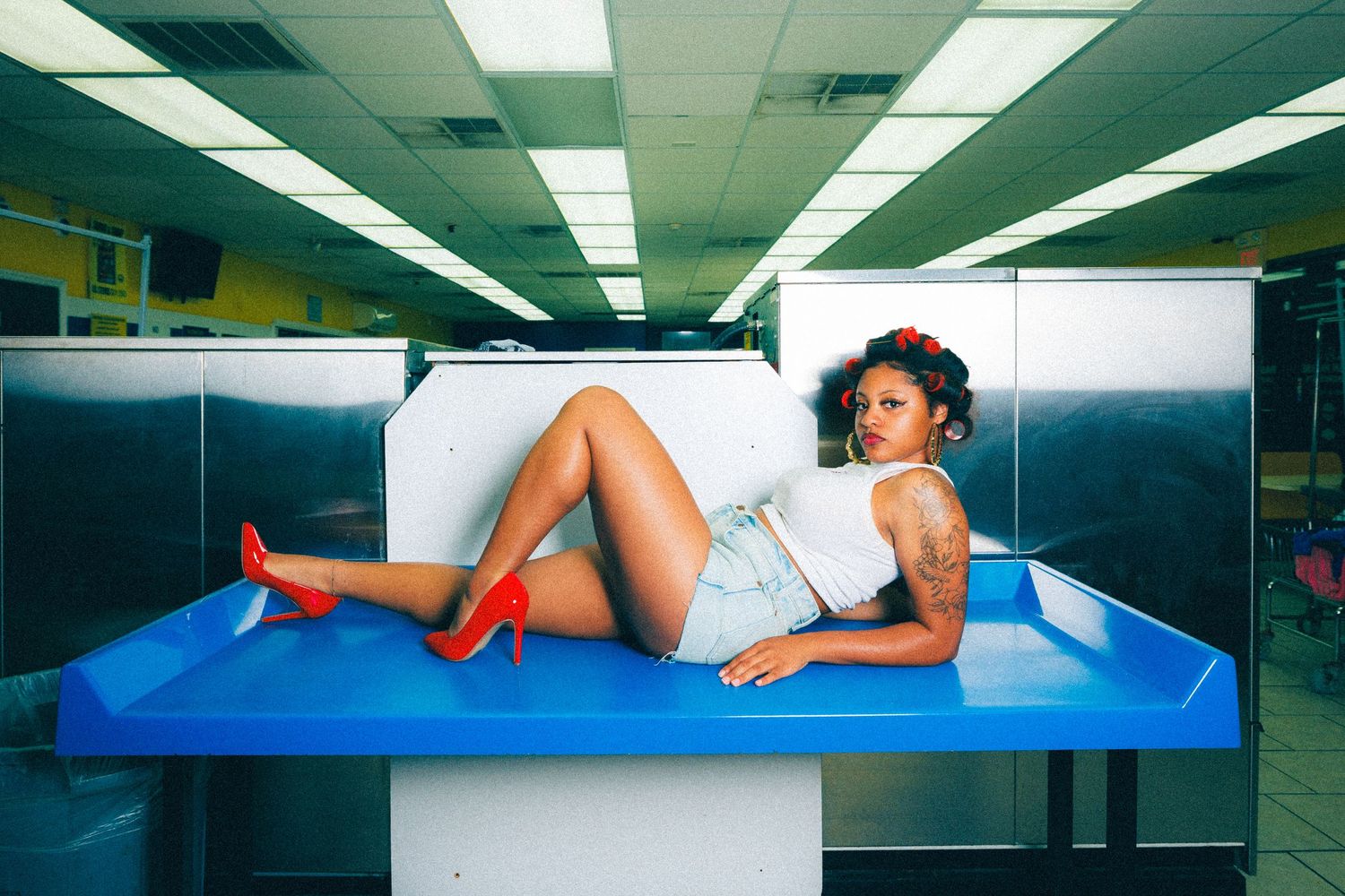 A tattooed woman in white crop top and denim shorts poses on a blue counter wearing red heels in a laundromat.