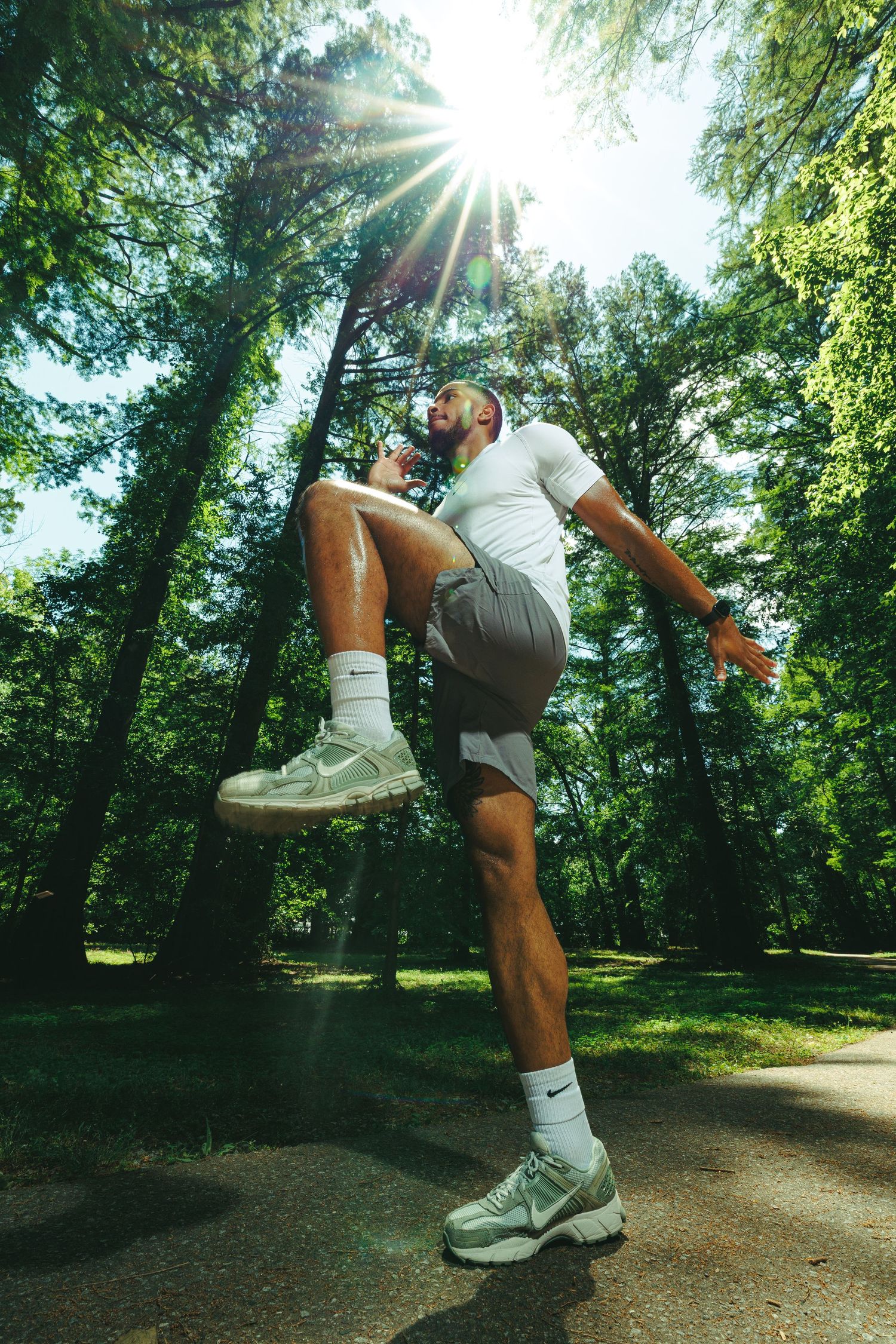 Athletic man in white shirt and gray shorts doing high knee exercise outdoors in a sunny green park.
