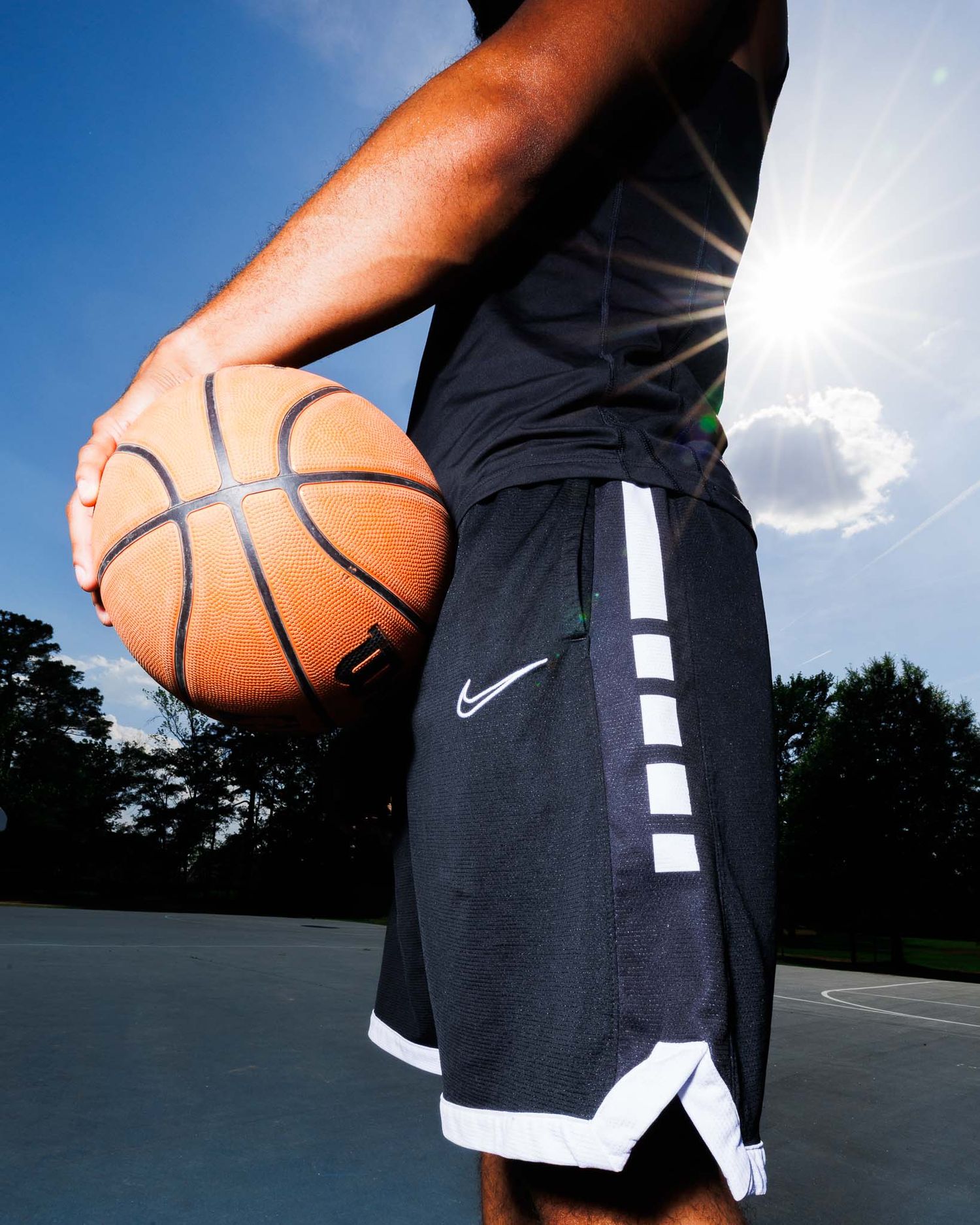 Basketball player in Nike Elite shorts holding a basketball outdoors on a court with bright sunlight.
