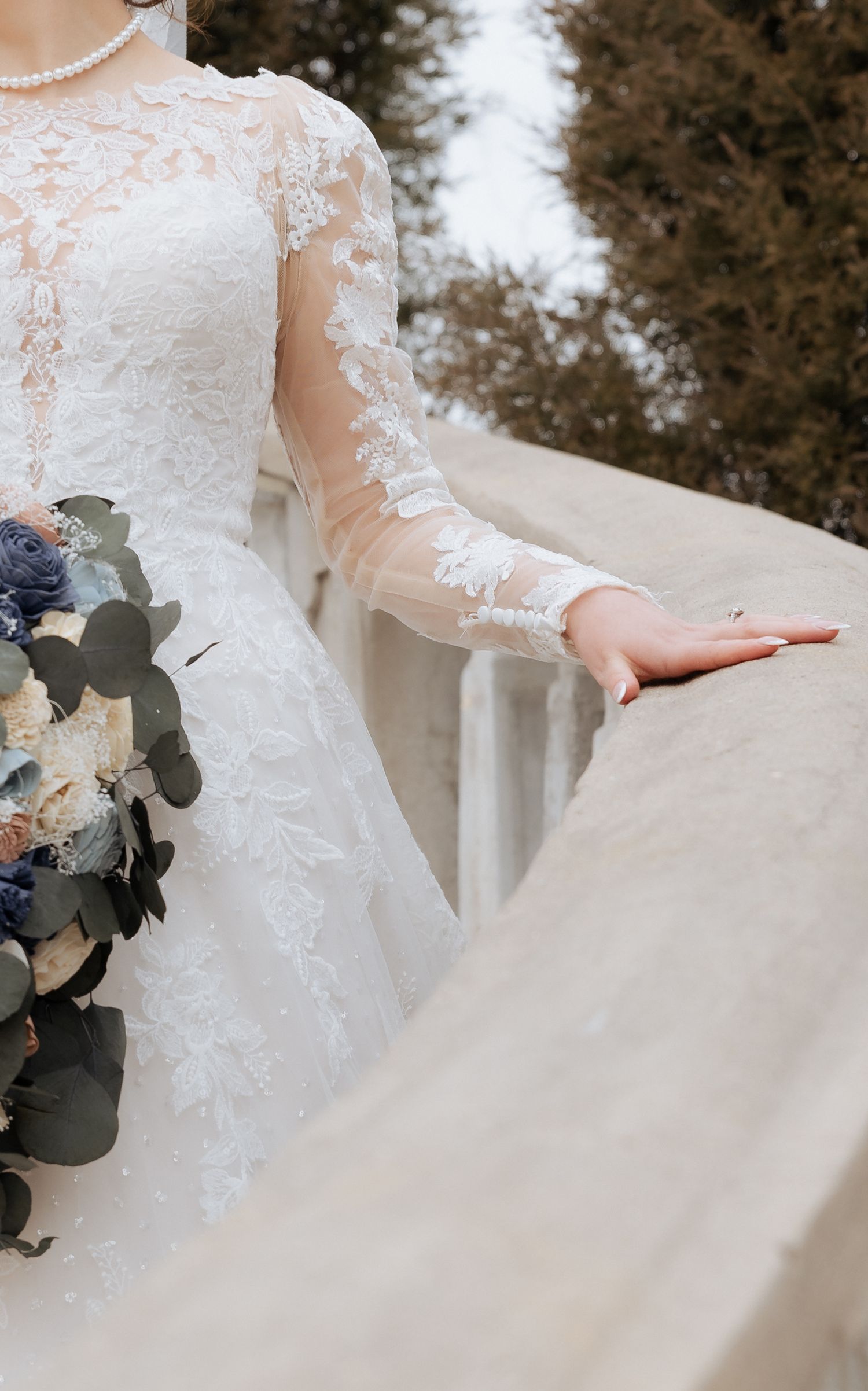 A bride in a delicate lace sleeve wedding gown holds a blue and white floral bouquet against snow-covered scenery.