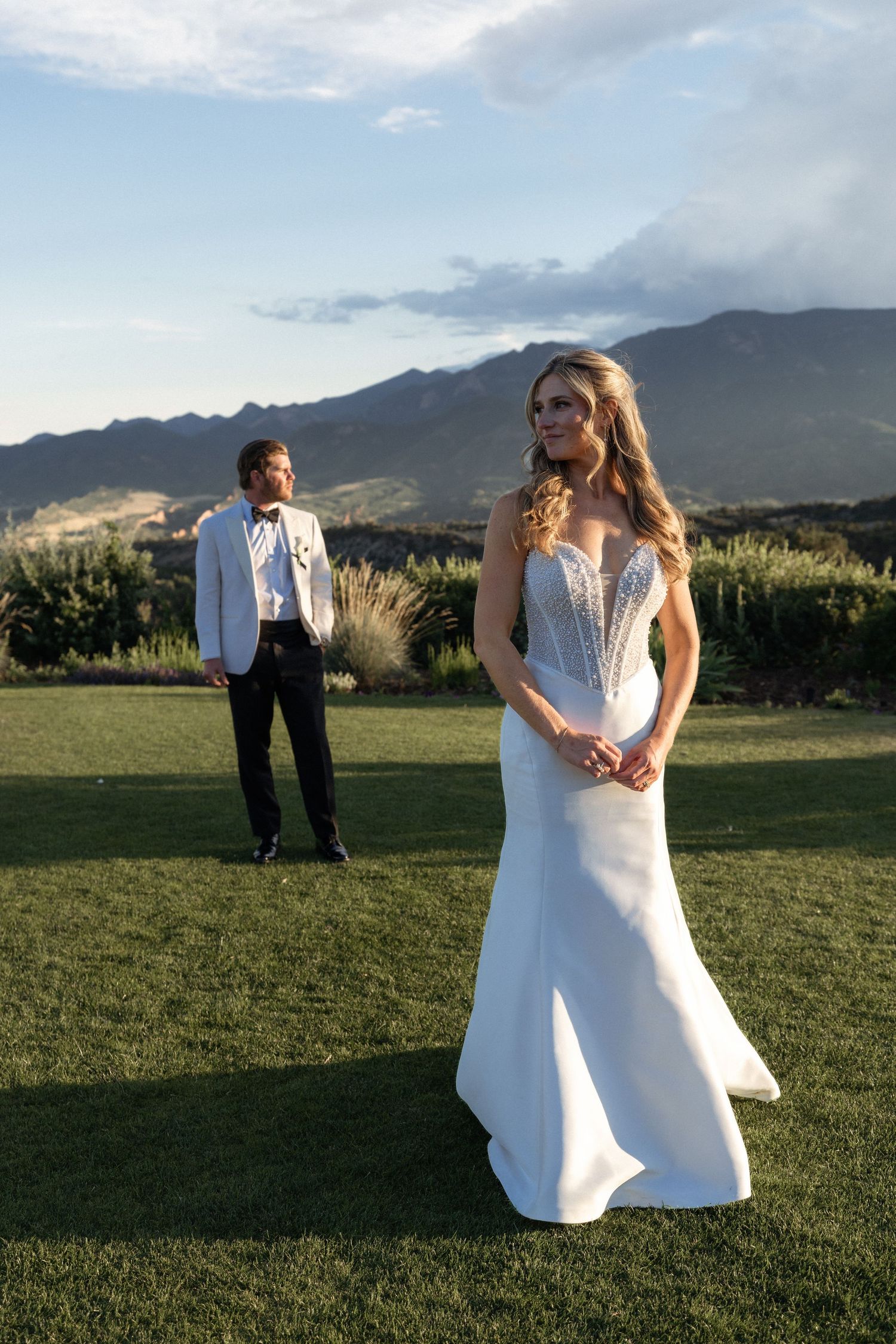 Bride in white gown stands on green lawn with groom in white jacket, mountains in background.