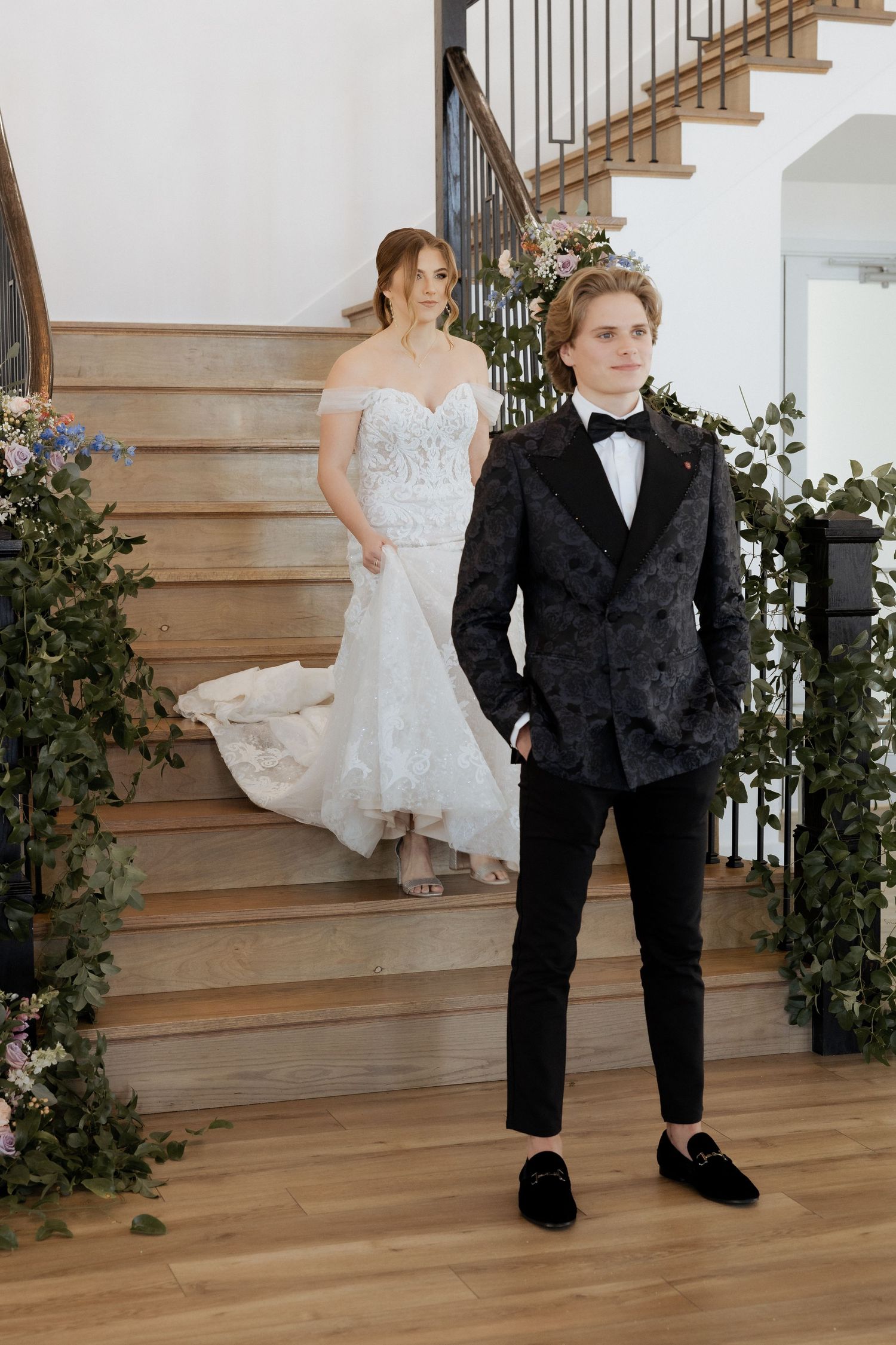 A formal wedding scene on a wooden staircase decorated with greenery, featuring elegant bridal attire and black tie formalwear.