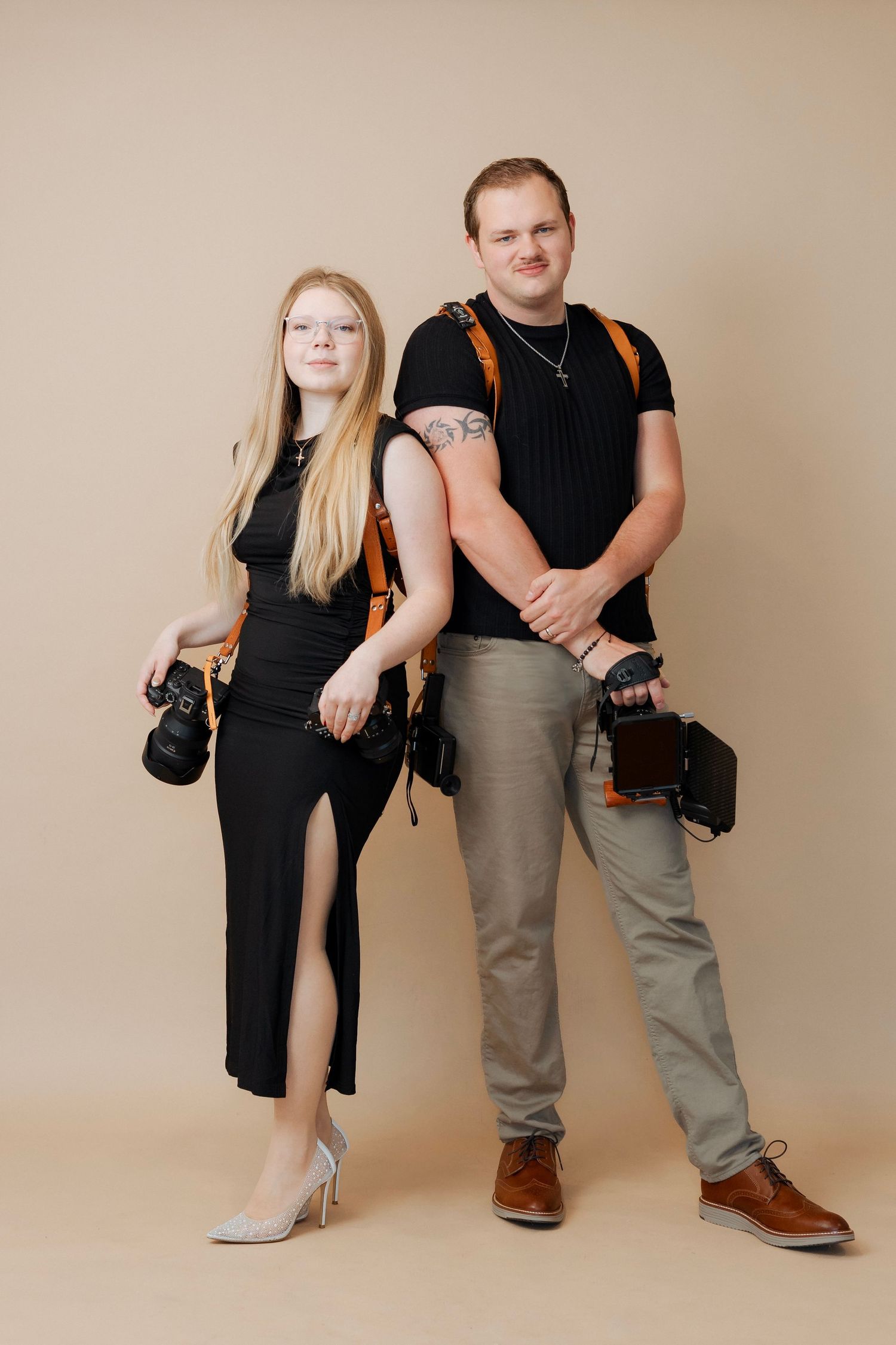 Professional photography partners in matching black outfits with camera gear posing against beige studio backdrop.