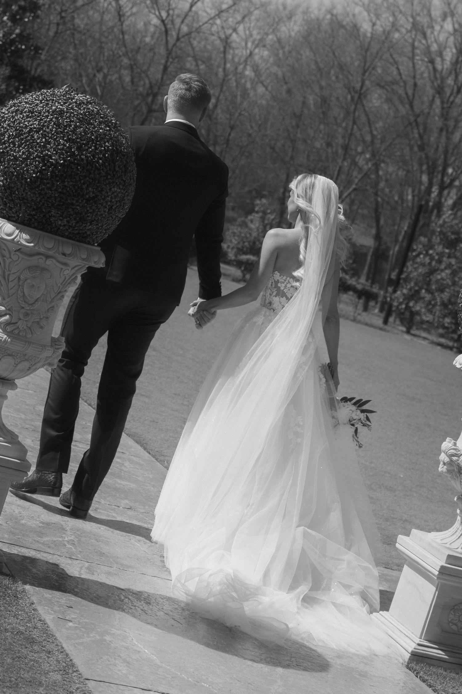 Black and white photo of bride and groom walking hand in hand through elegant garden grounds on their wedding day.