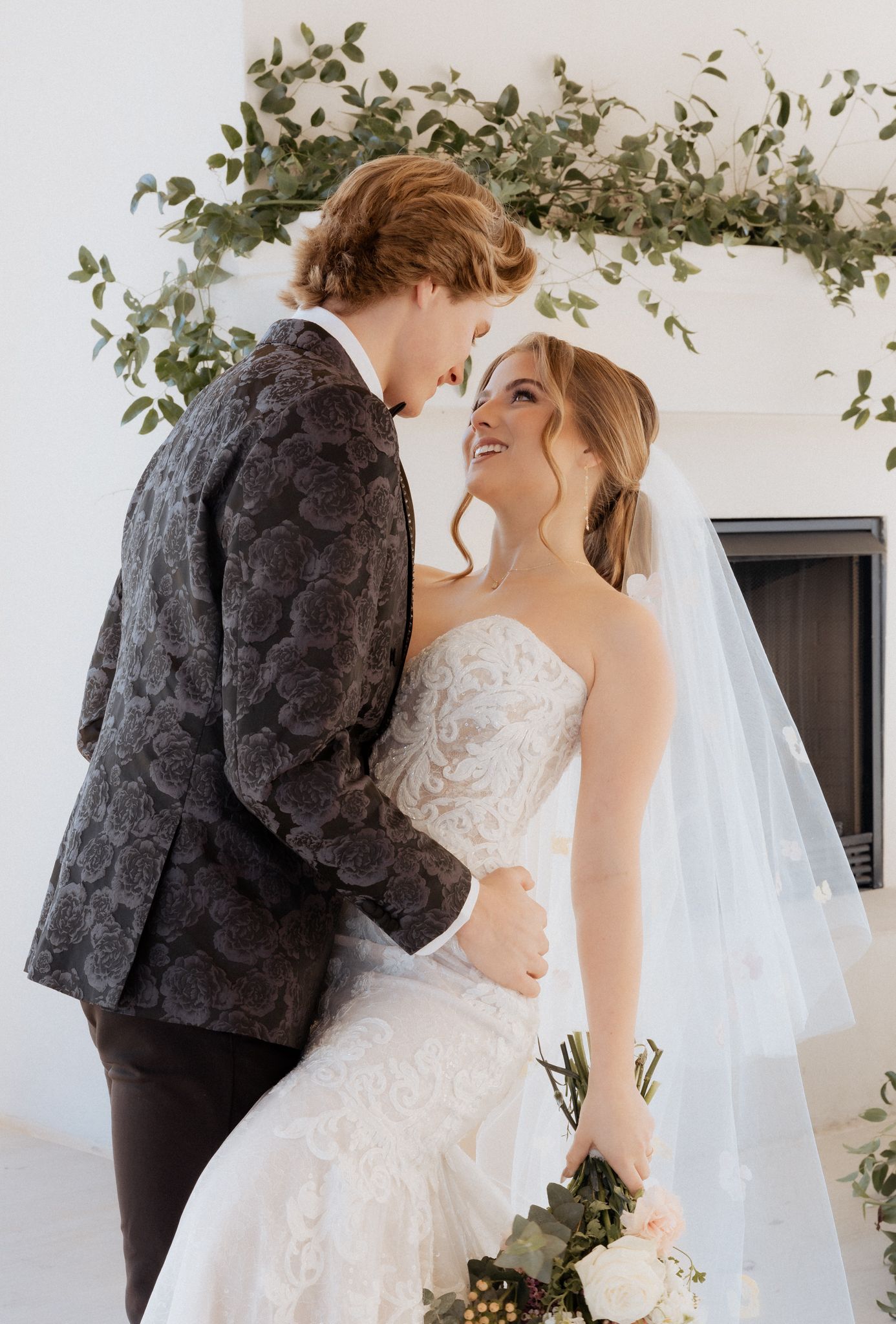 A romantic wedding moment between a couple under eucalyptus branch decor against a white wall.