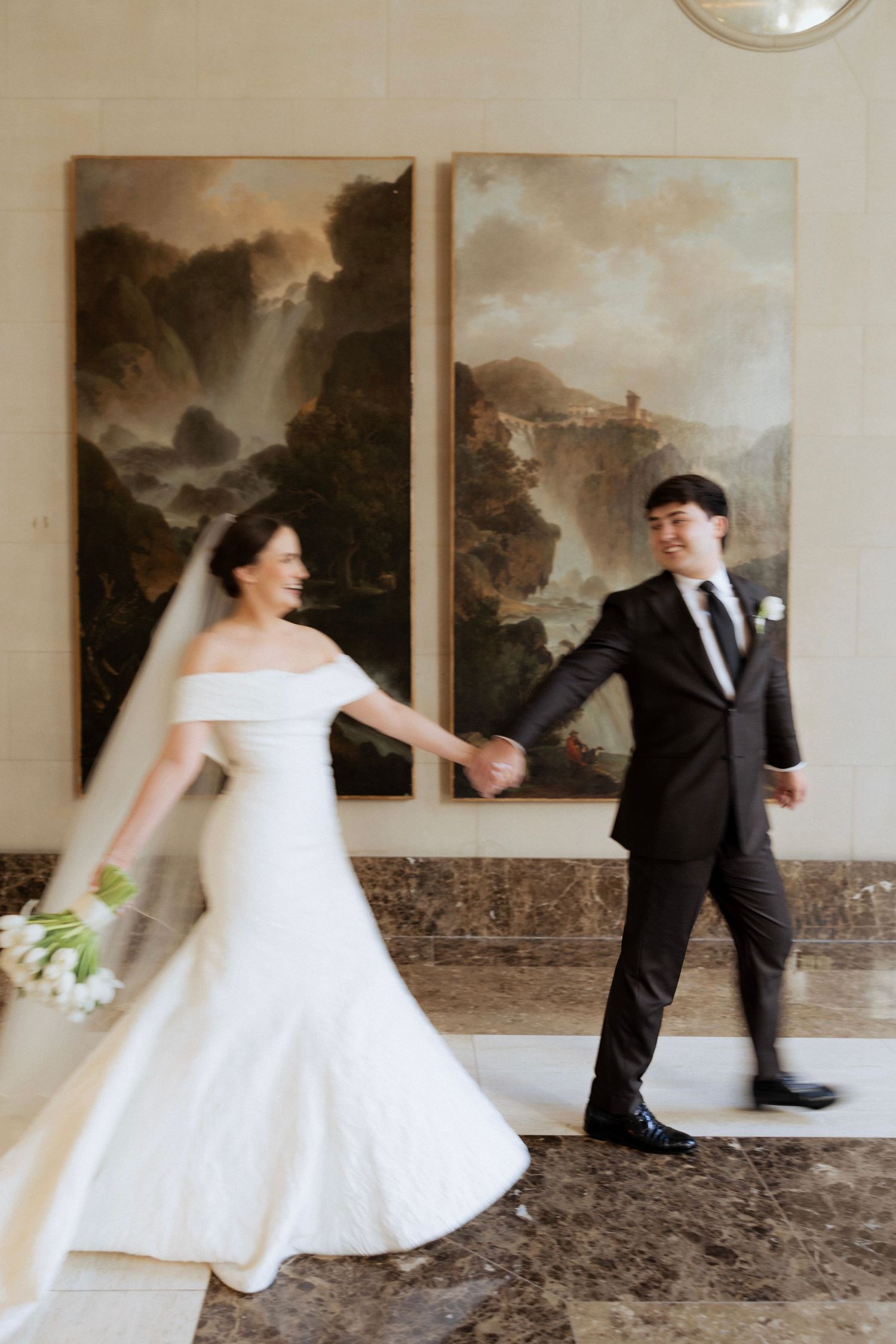 Bride in white off-shoulder gown holds hands with groom in black suit, walking past landscape paintings in elegant venue.