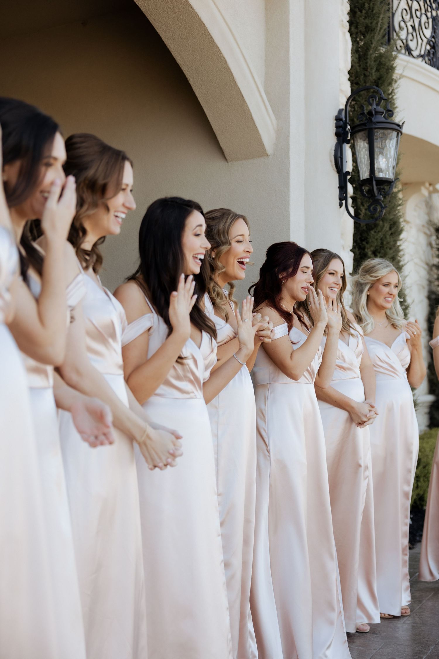 A row of bridesmaids in blush satin gowns laughing and clapping at an outdoor wedding venue.