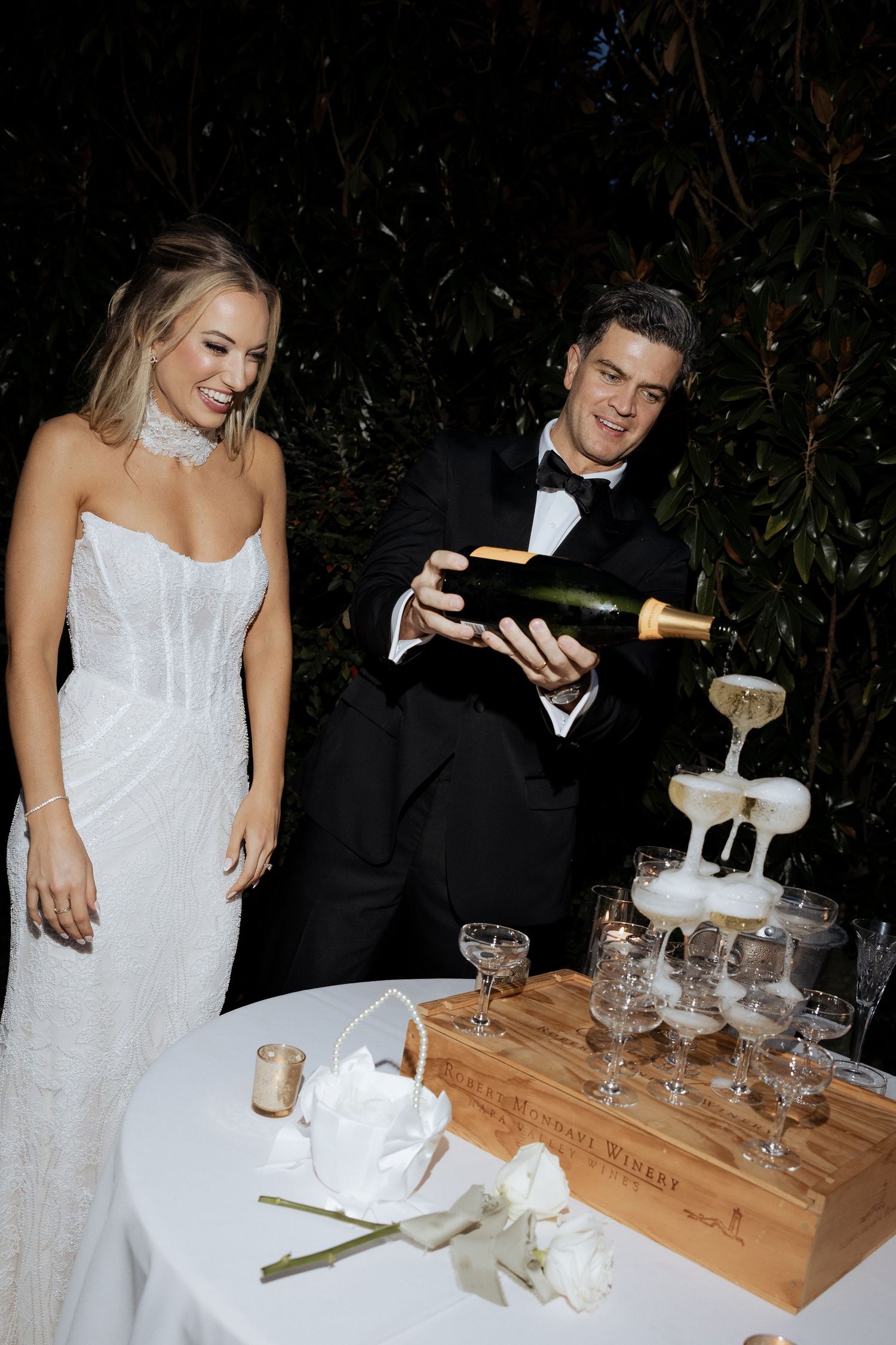A couple in formal attire pours champagne into a tower of glasses at a wedding celebration.