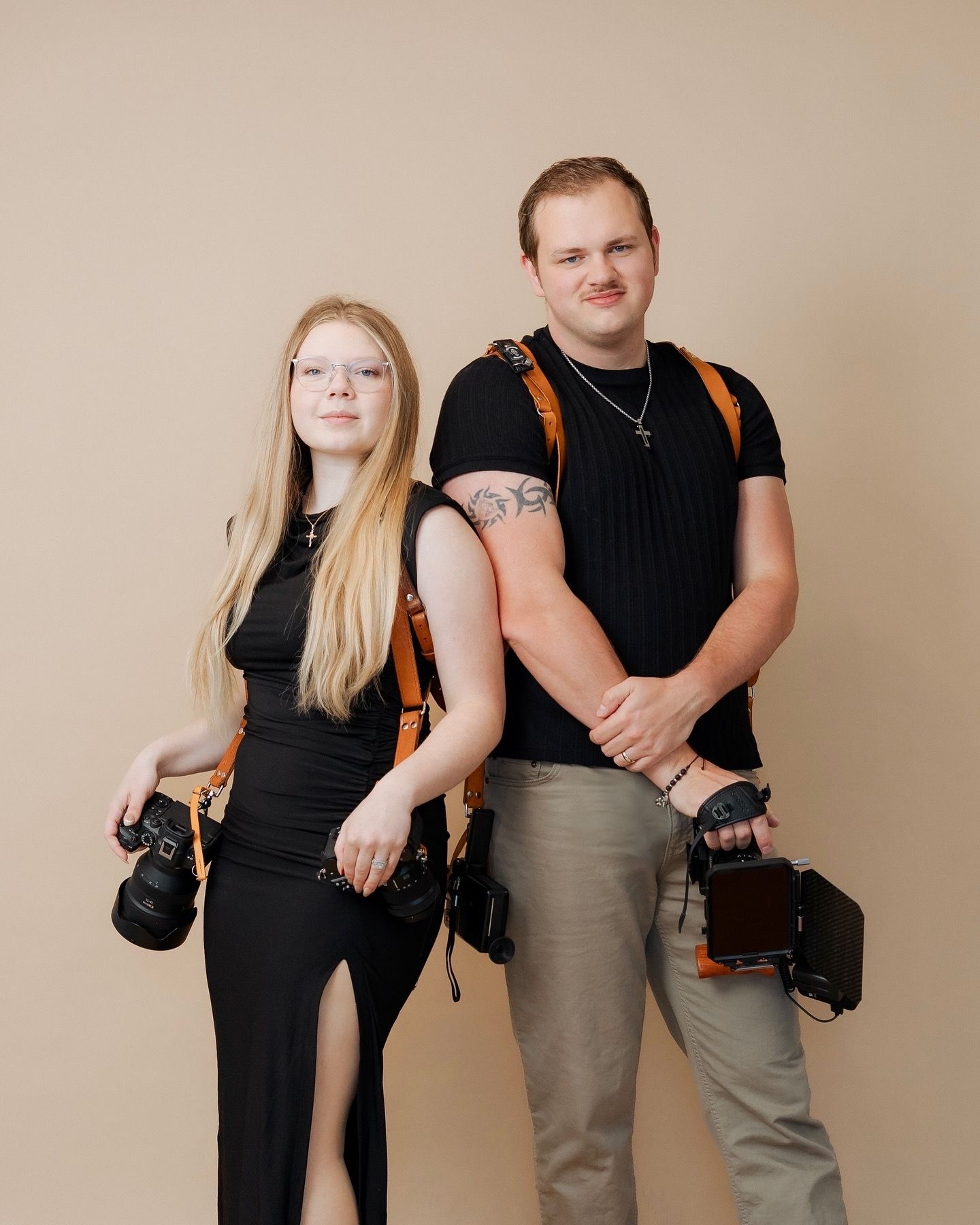 Two professional photographers wearing black tops stand with their cameras against a neutral background.