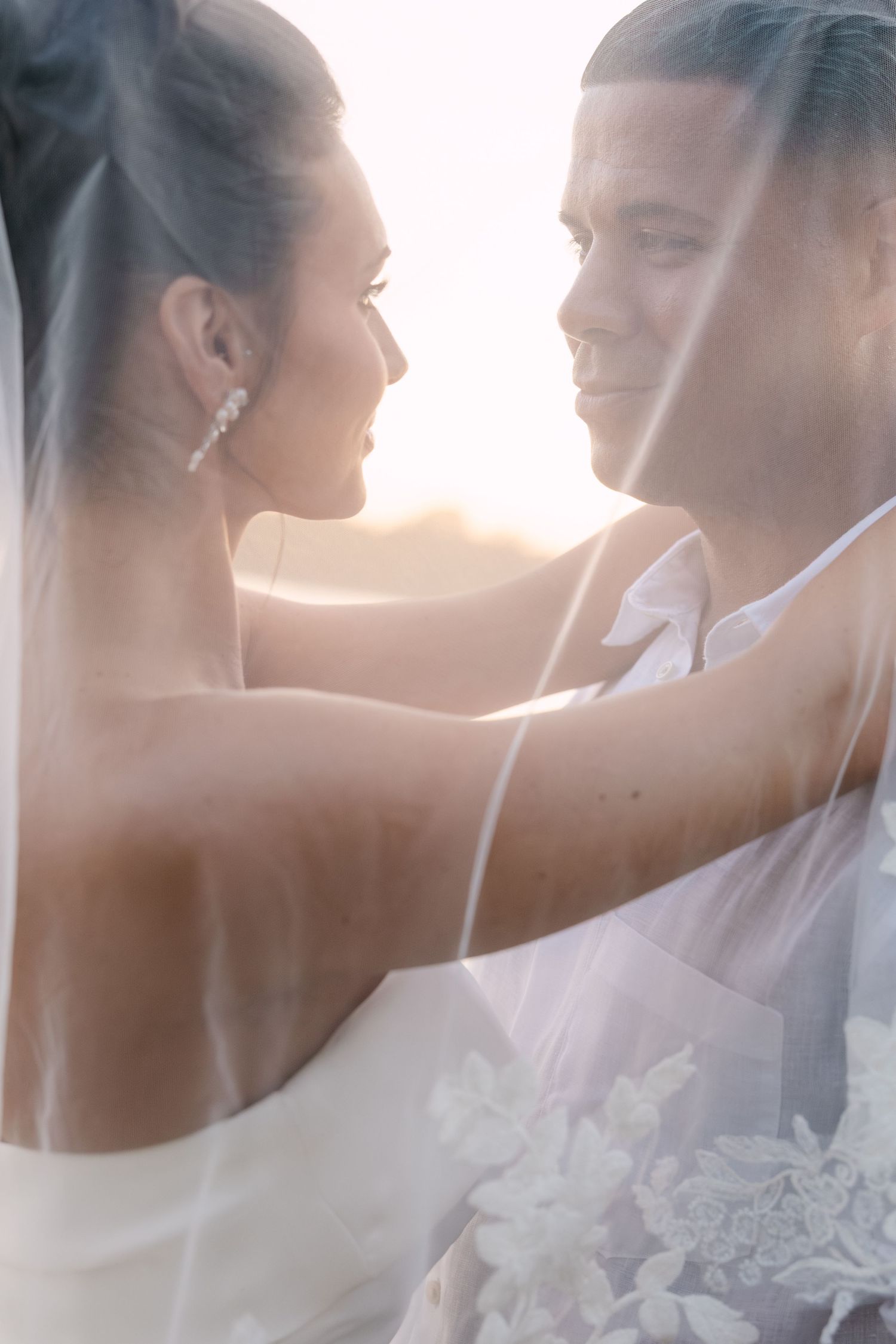 A romantic wedding couple embraces in soft sunlight through a delicate veil creating an ethereal moment.