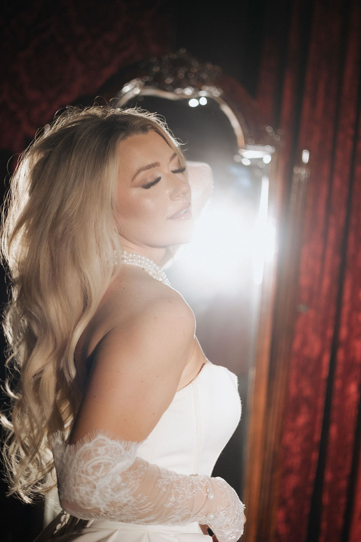 A bride in a white lace wedding dress poses in dramatic lighting against a red velvet curtain backdrop.